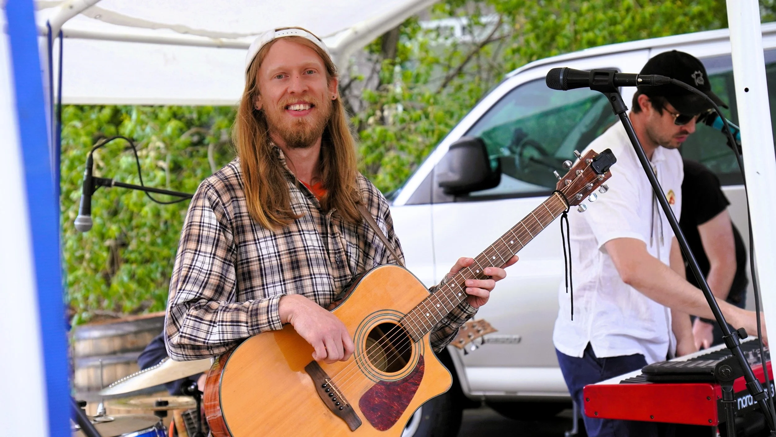 A man with long hair, a beard, and a plaid shirt, playing an acoustic guitar at an outdoor event, with another musician playing a keyboard in the background.