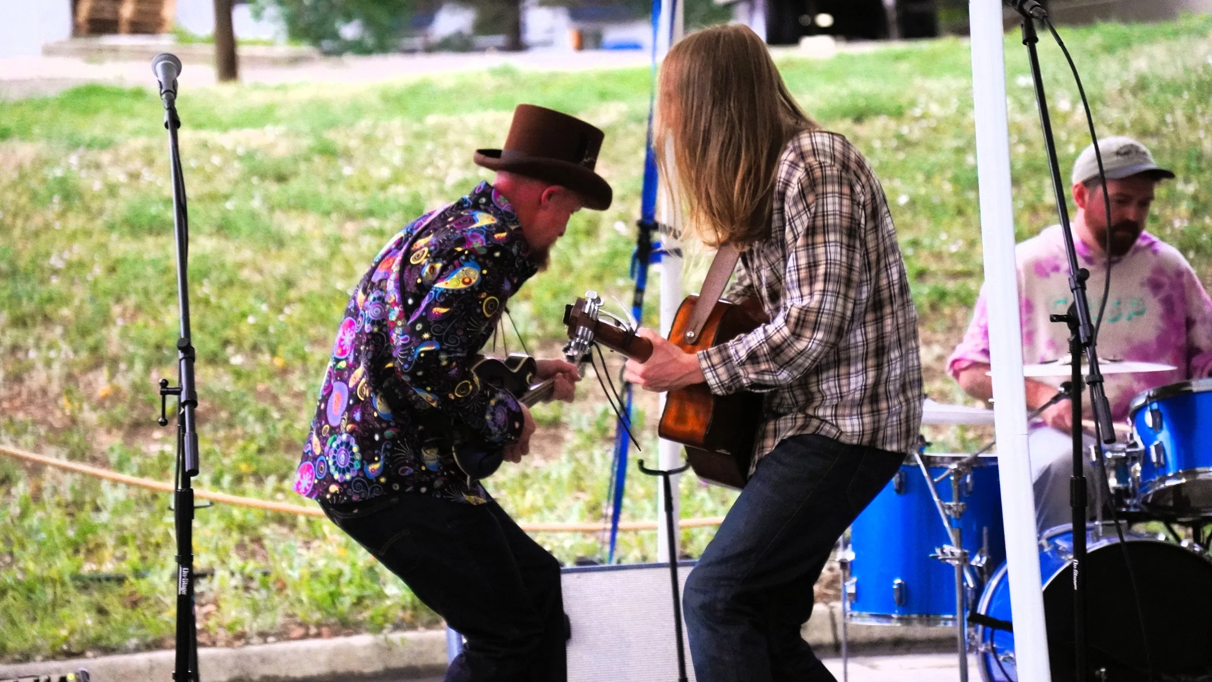 Two musicians on stage, one wearing a colorful shirt and a brown top hat playing a mandolin, and the other wearing a plaid shirt playing an acoustic guitar, with a drummer visible in the background.