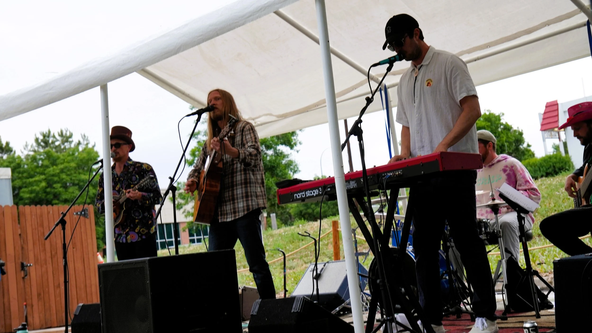 A band performing outdoors under a canopy, with musicians playing guitar, keyboard, and drums.