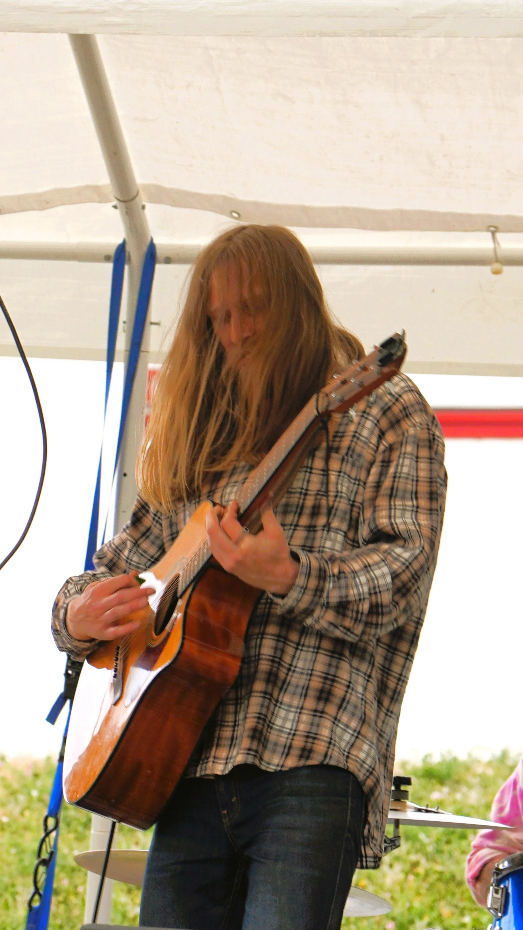 A person with long hair playing an acoustic guitar under a tent or canopy, wearing a plaid shirt, with a drum set partially visible in the background.