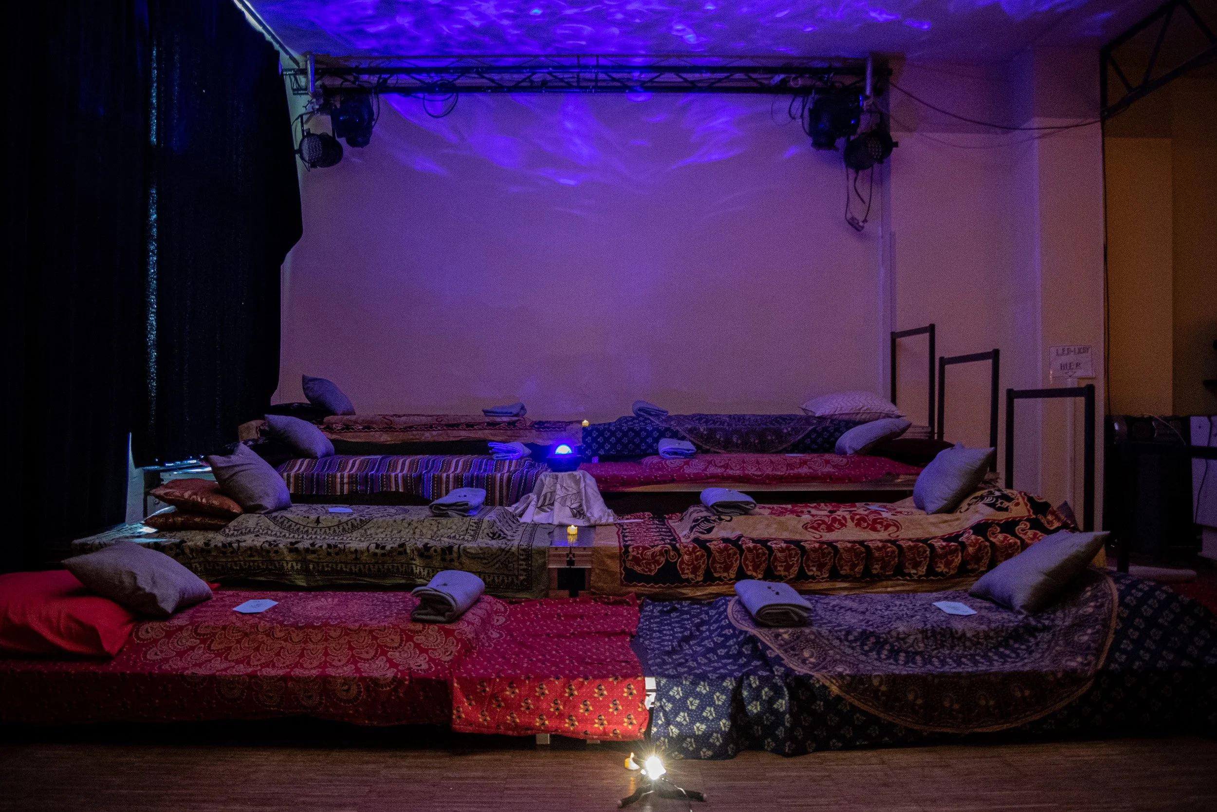 Soul Spa Berlin Live Theta Traumreise. Ein Raum mit mehreren aufgestellten Matratzen und Kissen, gedeckten Tücher und kleine Kerzen, mit blauer Beleuchtung und dunklen Vorhängen im Hintergrund.