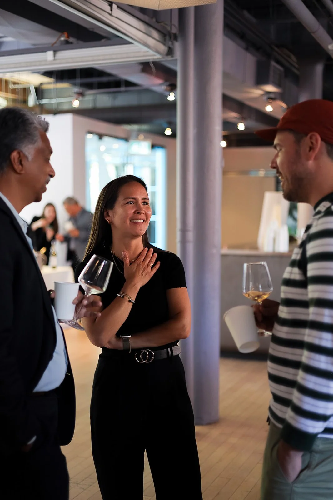 Three people engaged in conversation at a social gathering, holding wine glasses and cups in a modern indoor setting.