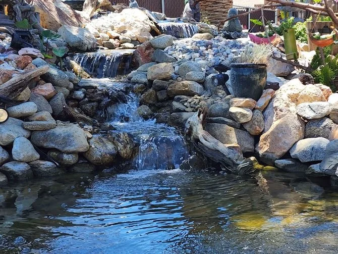 A small artificial waterfall flowing into a pond, surrounded by rocks and plants, with decorative elements like a planter and bamboo in the background.