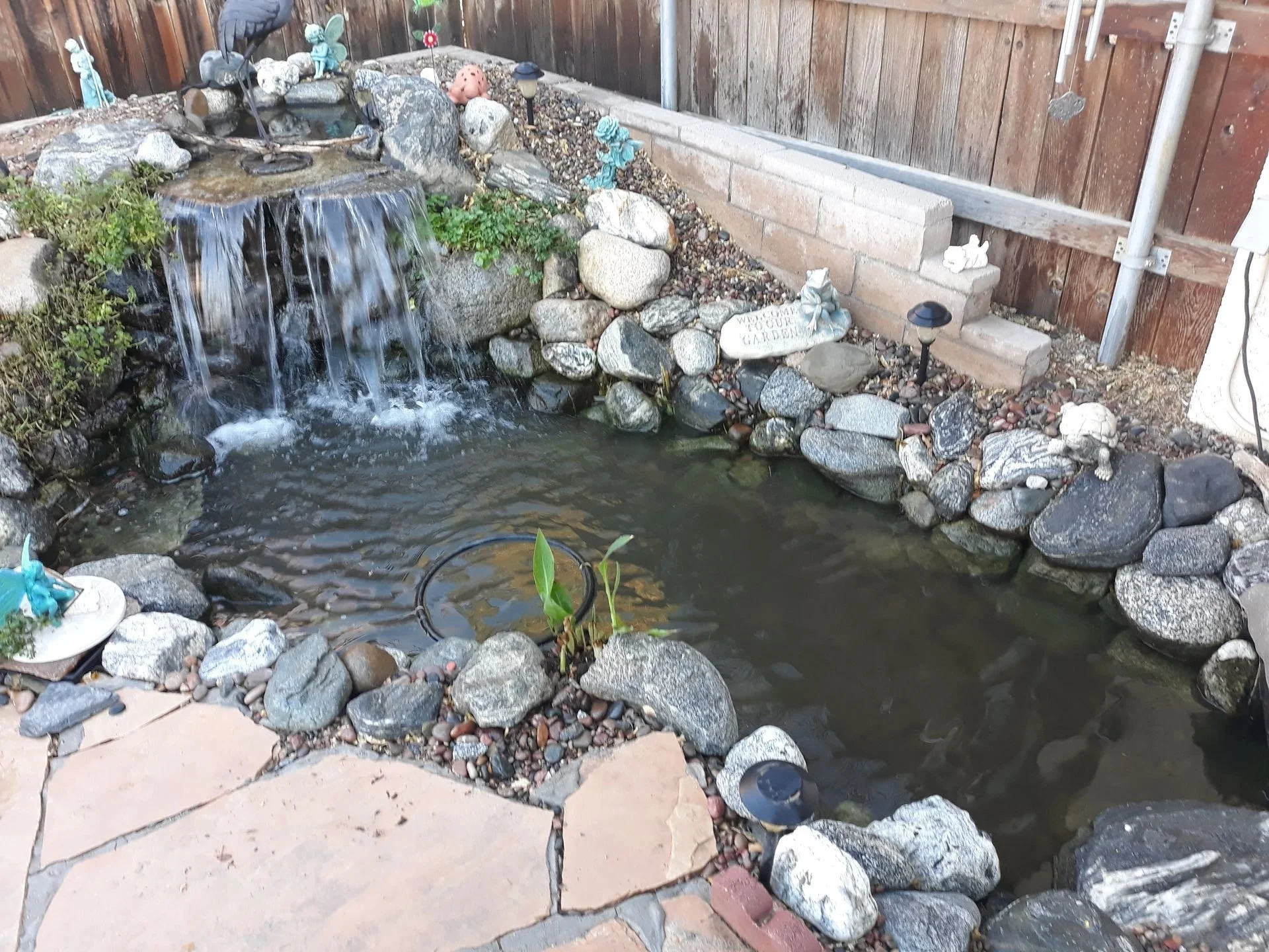 Small backyard pond with waterfall feature surrounded by rocks, plants, decorative garden ornaments, and solar-powered garden lights, enclosed by a wooden fence and brick wall.