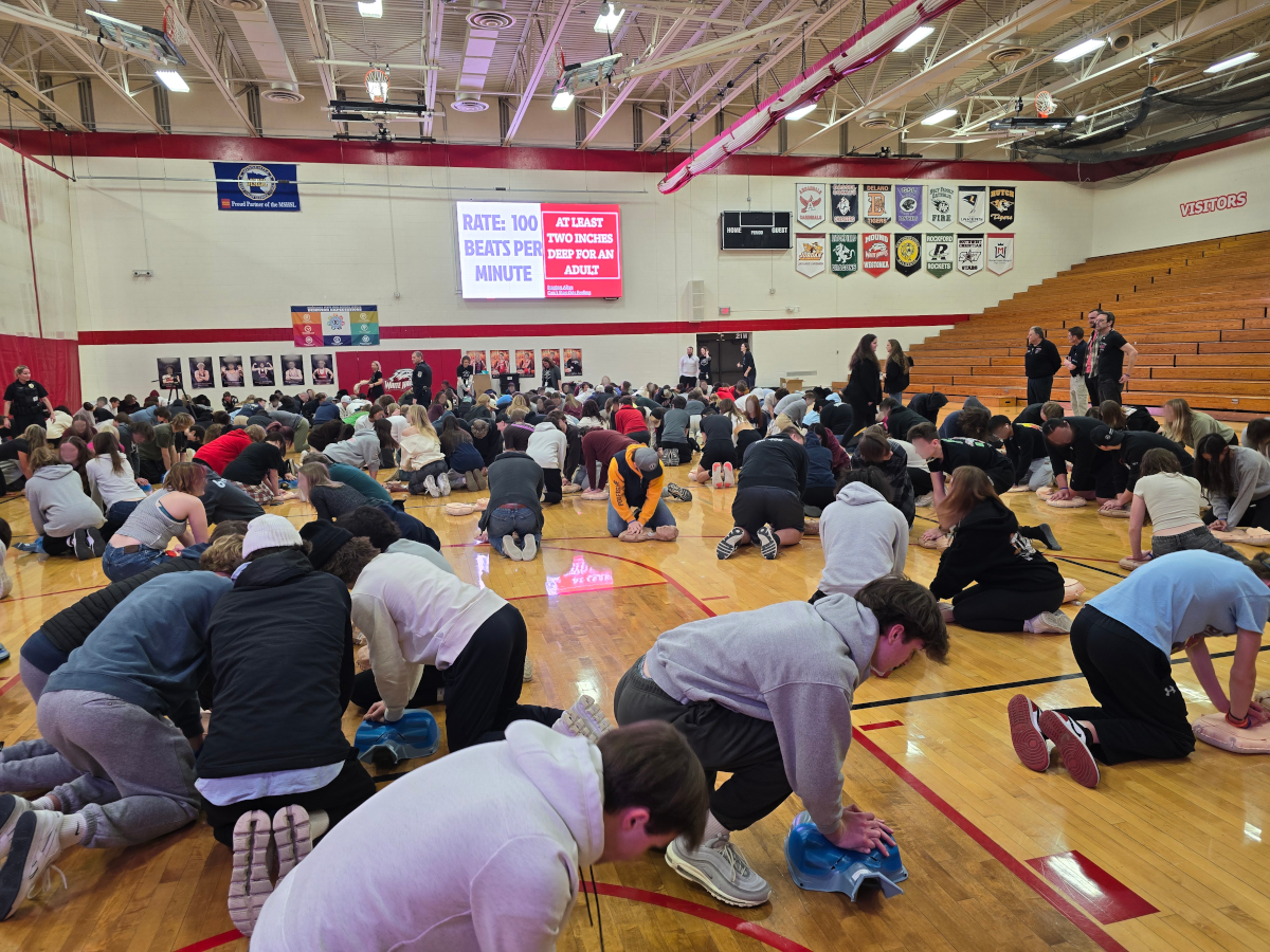 A large group of students practicing CPR in a gymnasium during a  HeartSafe CPR training session, with an instructor at the front and banners on the walls.
