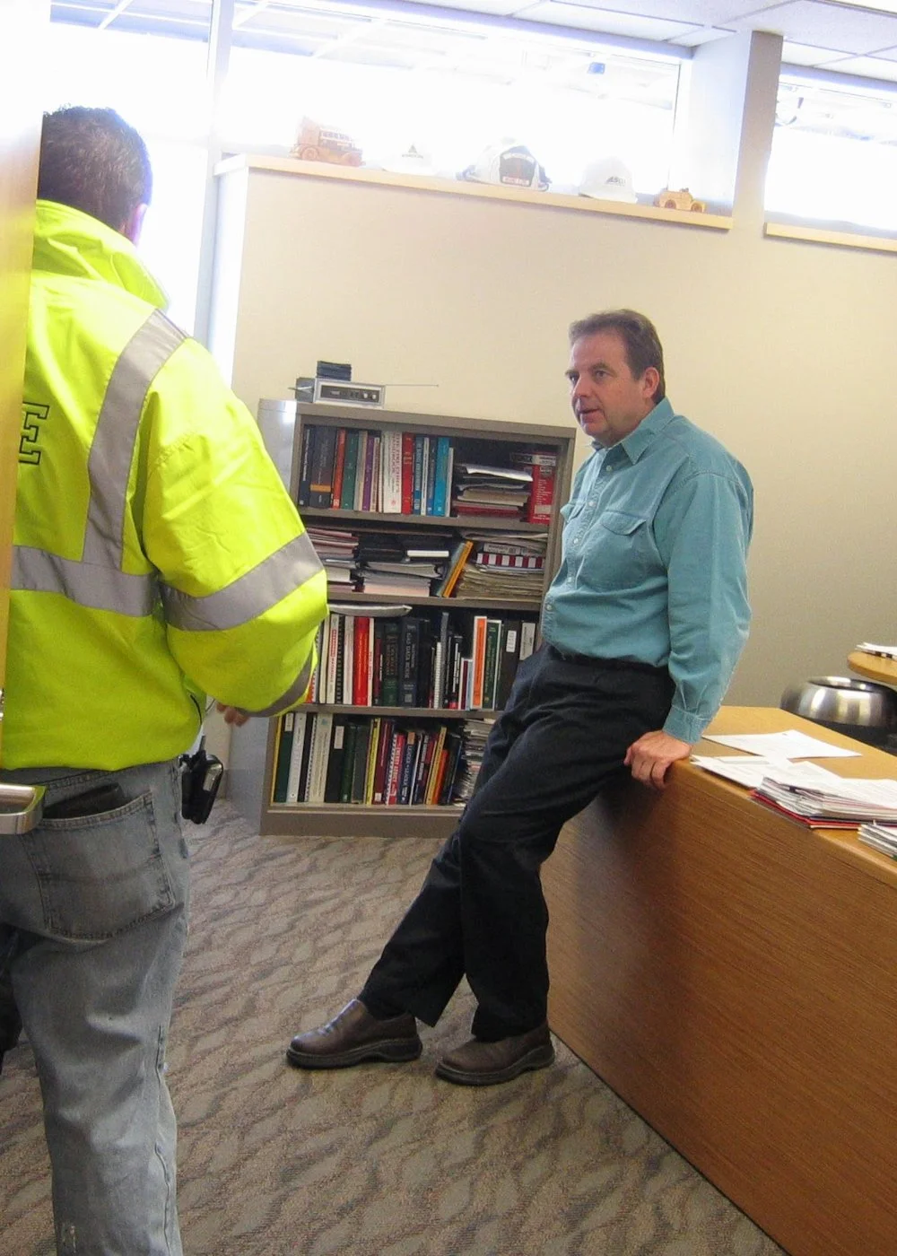 A Mound city employee and Greg Pederson having a conversation in an office, one in a high-visibility jacket facing away and Greg in business casual attire leaning against a wooden desk.