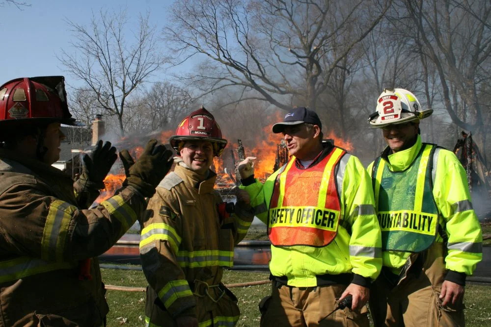 Four firefighters at a training burn in front of a house on fire, with flames and smoke. Greg Pederson acts as safety officer and converses with firefighters participating.