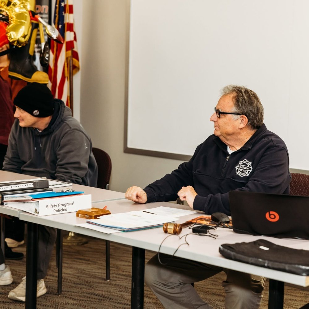 Greg Pederson and fire fighter Heitz sitting at a table with documents, a gavel, and electronic devices in a meeting room.