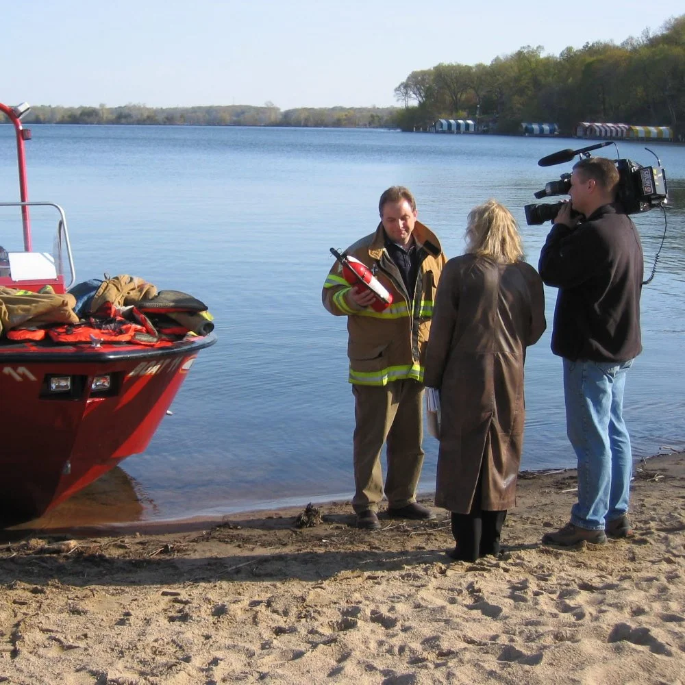 A news crew interviews a Greg Pederson at Surfside Beach in Mound, MN.  They stand next to a red rescue boat, with a calm lake and trees in the background.