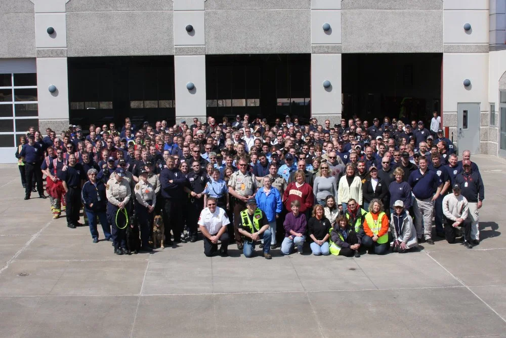 Large group of firefighters and emergency responders gathered outdoors in front of a fire station for a group photo.