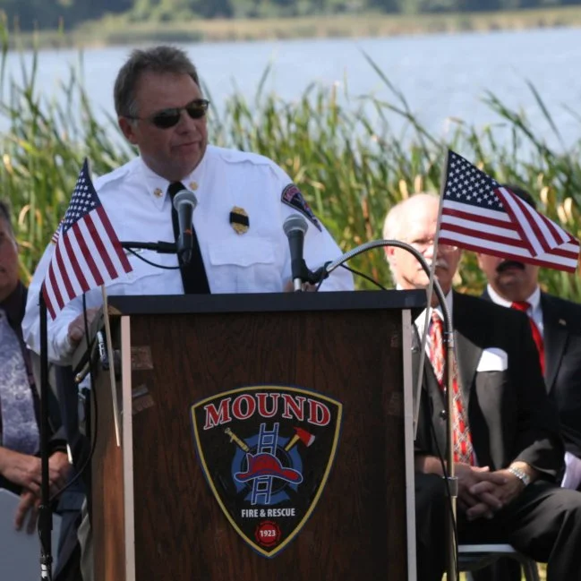 Greg Pederson in fire department uniform giving a speech at an outdoor event, with American flags on either side of the podium, set against a lakeside background.