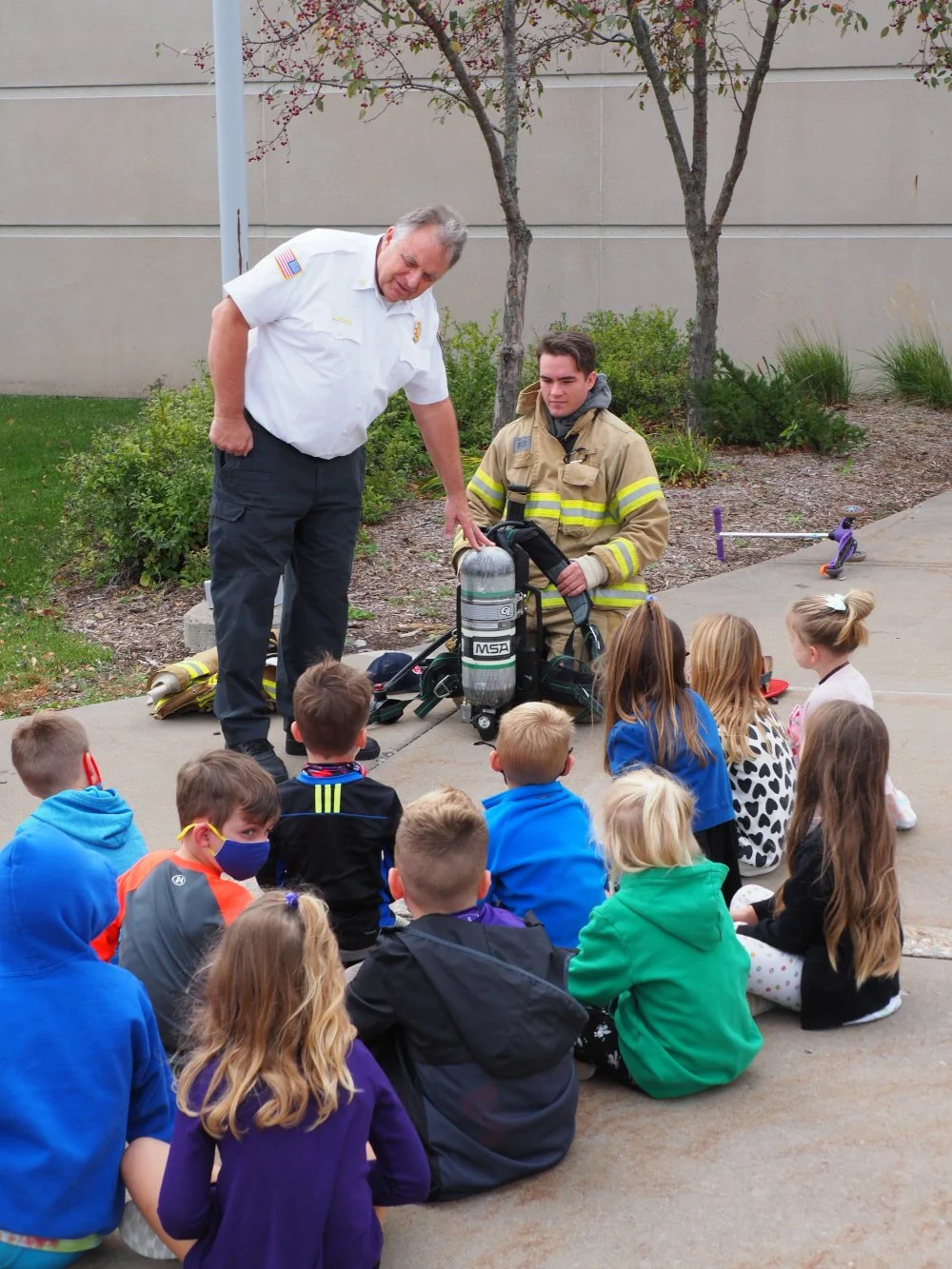 Firefighter Greg Pederson and firefighter Palm giving a safety presentation to a group of children sitting on the sidewalk outside a school.