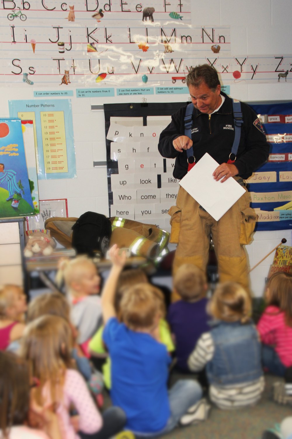 Greg Pederson in firefighter gear giving a safety talk to a group of young children in a classroom, with educational posters and the alphabet on the wall behind him.