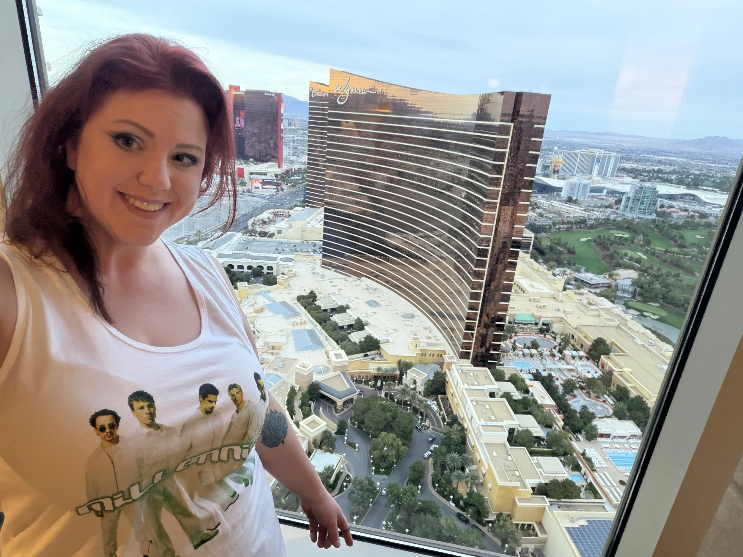 Woman with red hair smiling inside a tall building, with a view of Las Vegas skyline and hotels including the Wynn and Encore.