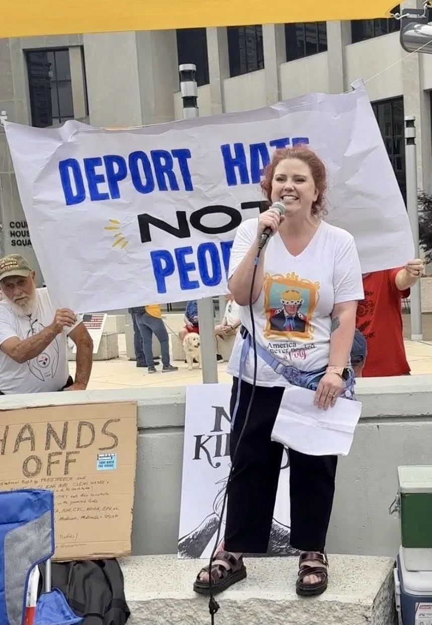 A woman speaking into a microphone at a protest rally with a sign behind her that reads 'Deport Hate, Not People'.