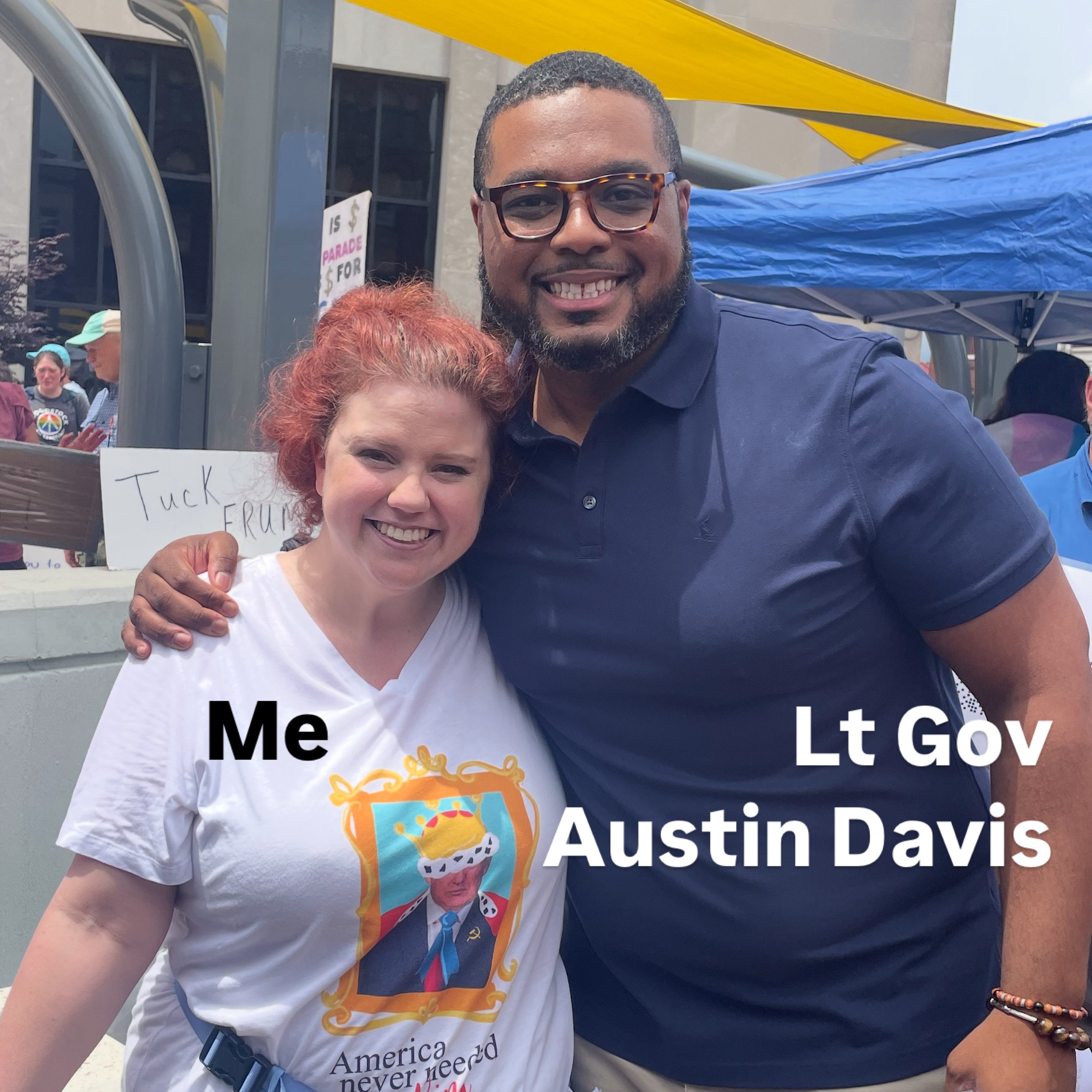 A smiling man with glasses and a beard standing next to a woman with red hair at a public event with tents in the background. The woman wears a white T-shirt with a design related to Austin, Texas. The image appears to be taken outdoors during the day.