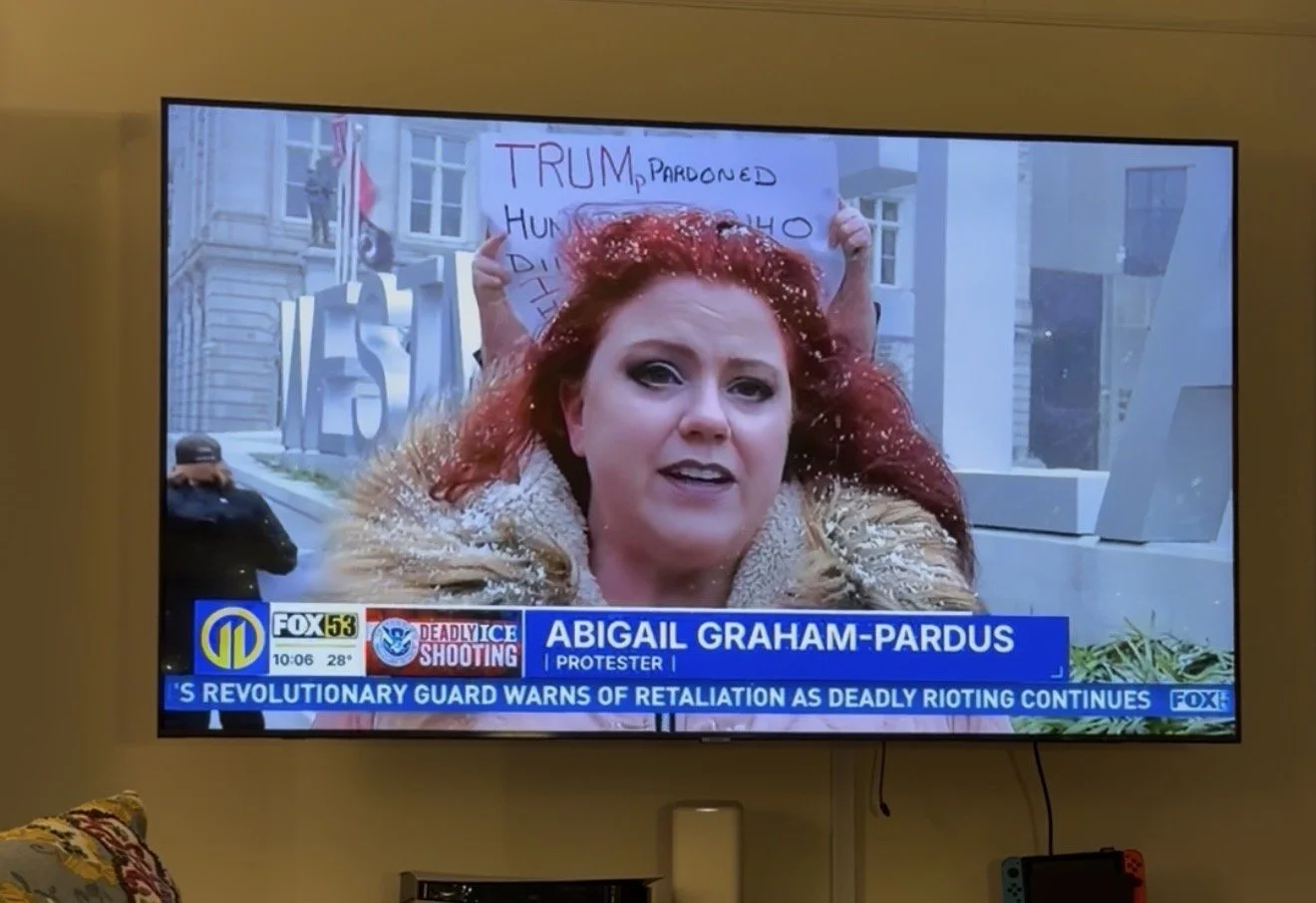 News screenshot showing a woman with red hair speaking on television, with a protest sign in the background, and news headlines about a deadly shooting and protests.