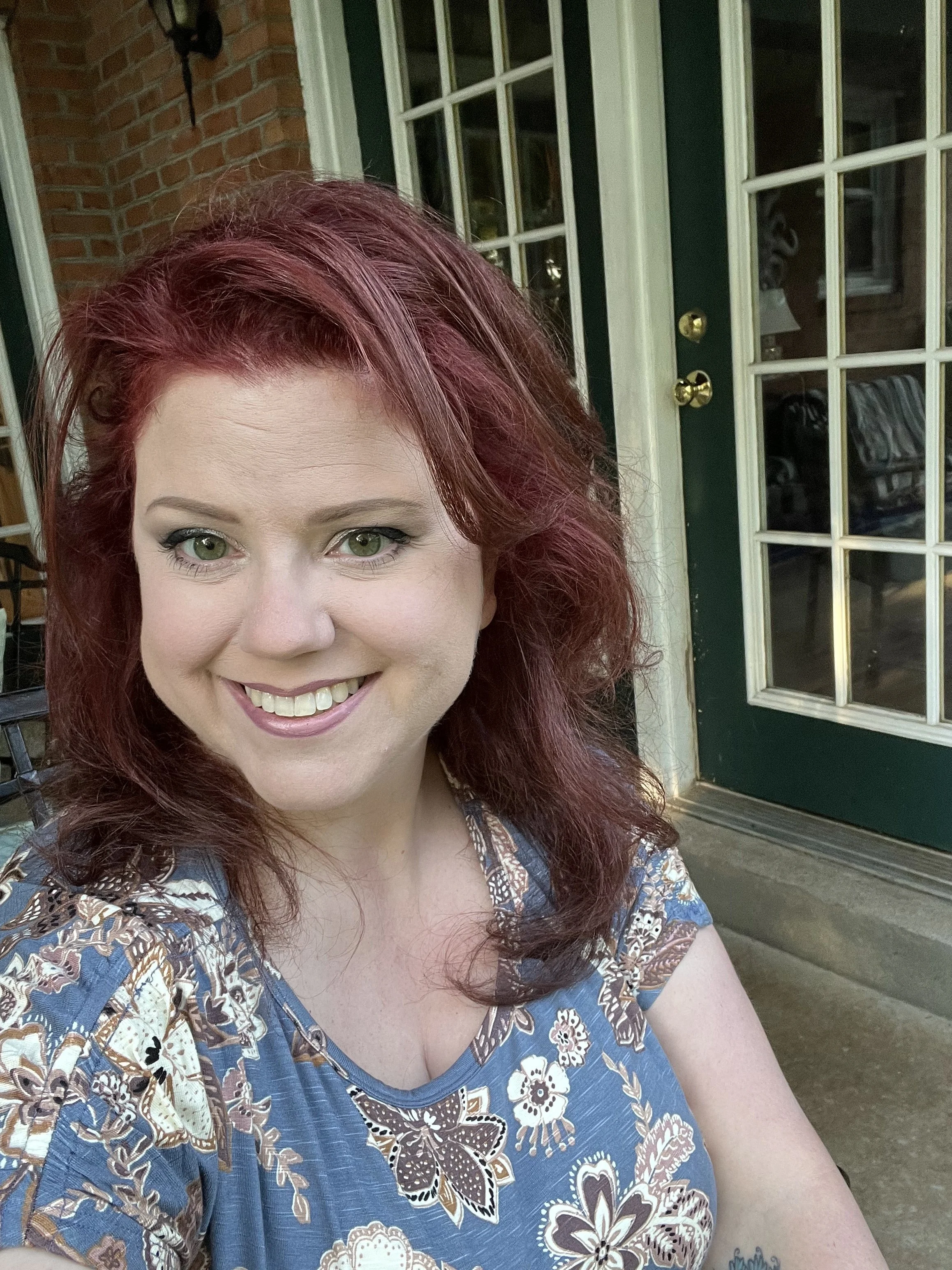 A woman with red hair and green eyes smiling outdoors in front of a glass door with a brick wall and porch area.