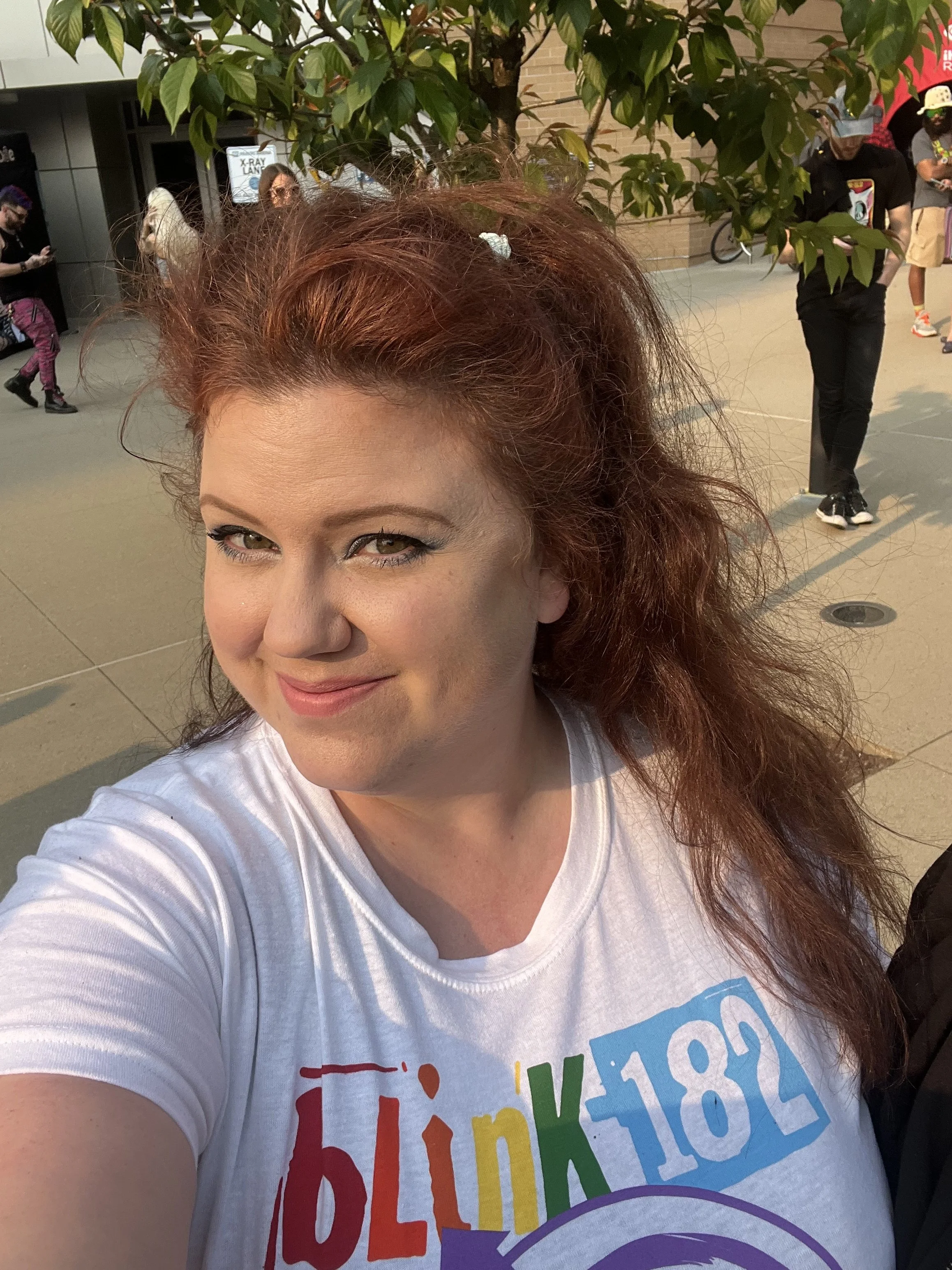 A woman with red hair and makeup posing for a selfie with a group of diverse people in the background outdoors near a brick building and a tree.