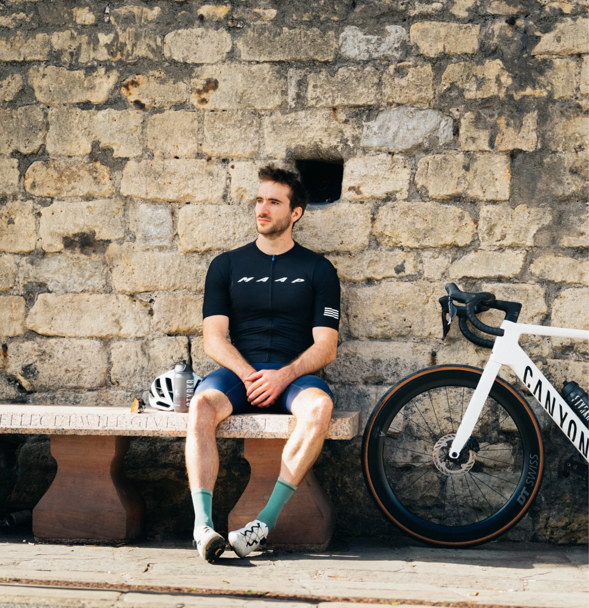 A young man with dark hair and a beard sitting against a large stone wall, wearing a black cycling shirt, shorts, and cycling shoes. A bicycle is parked next to him, and a white helmet and water bottle are on a bench in front of him.