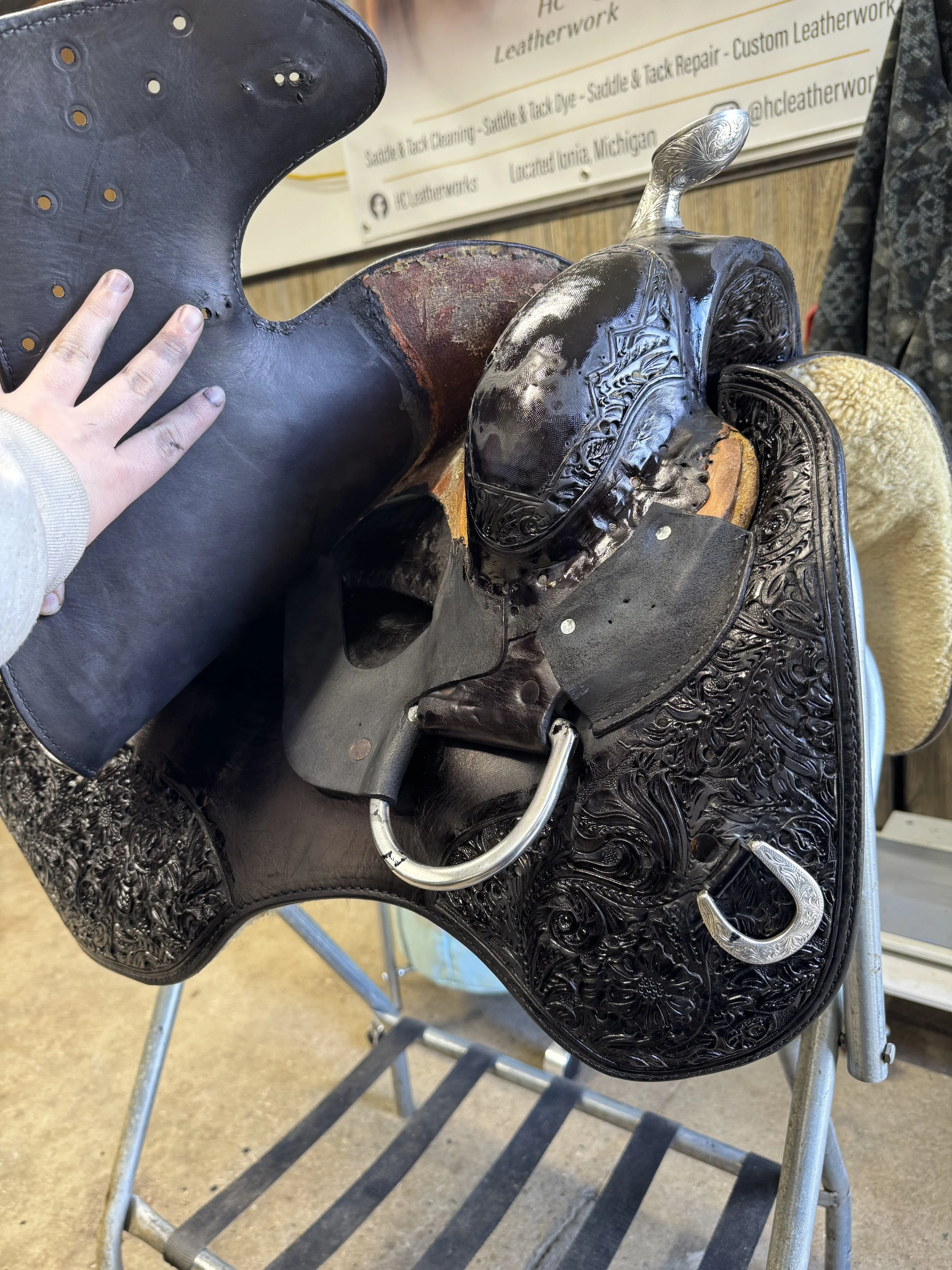 A black leather Western saddle on a saddle stand, with intricate floral engravings, in a tack room with a sign in the background.