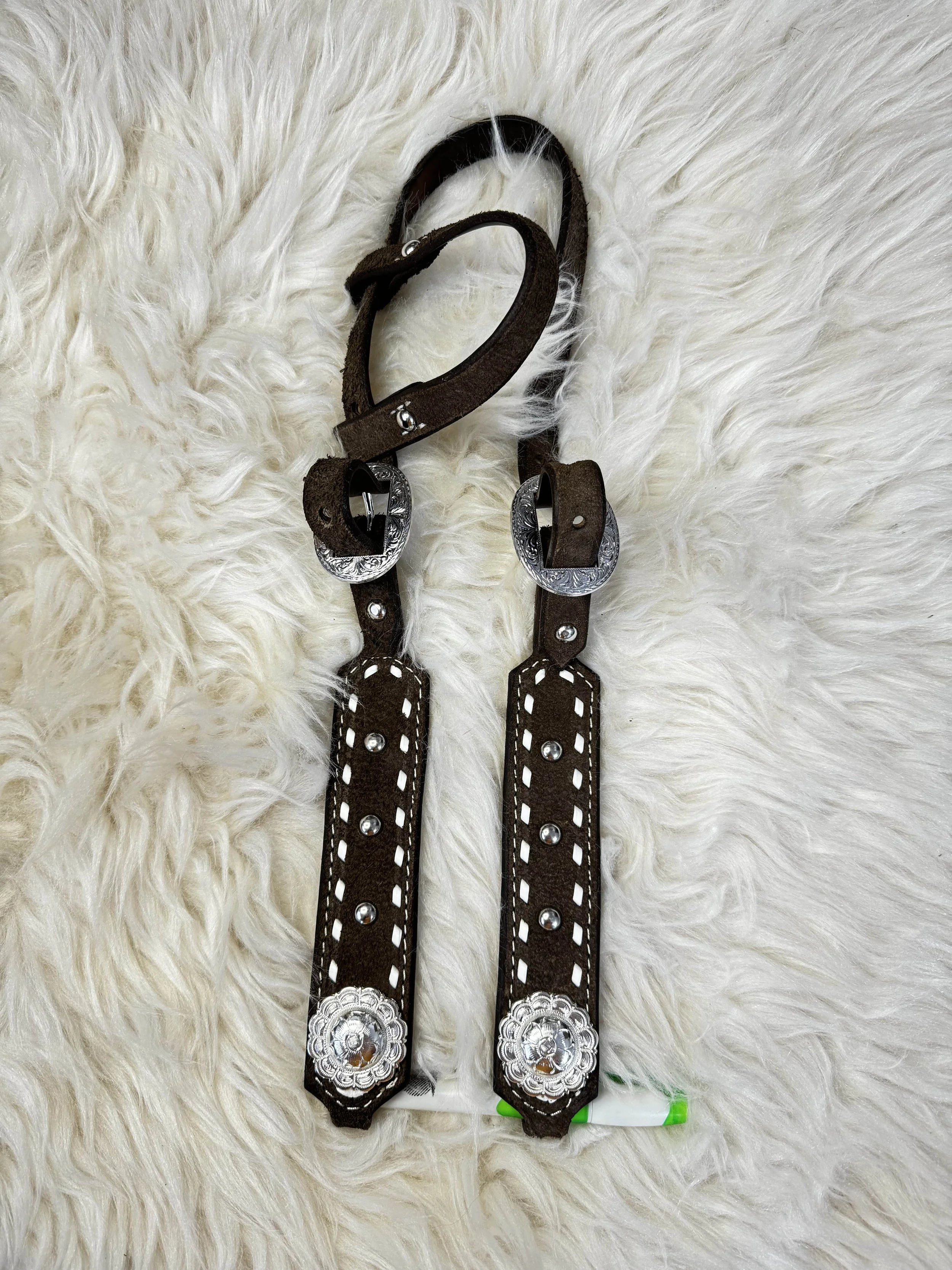 Two brown leather straps with silver decorative conchos and white stitching on a fluffy white surface.