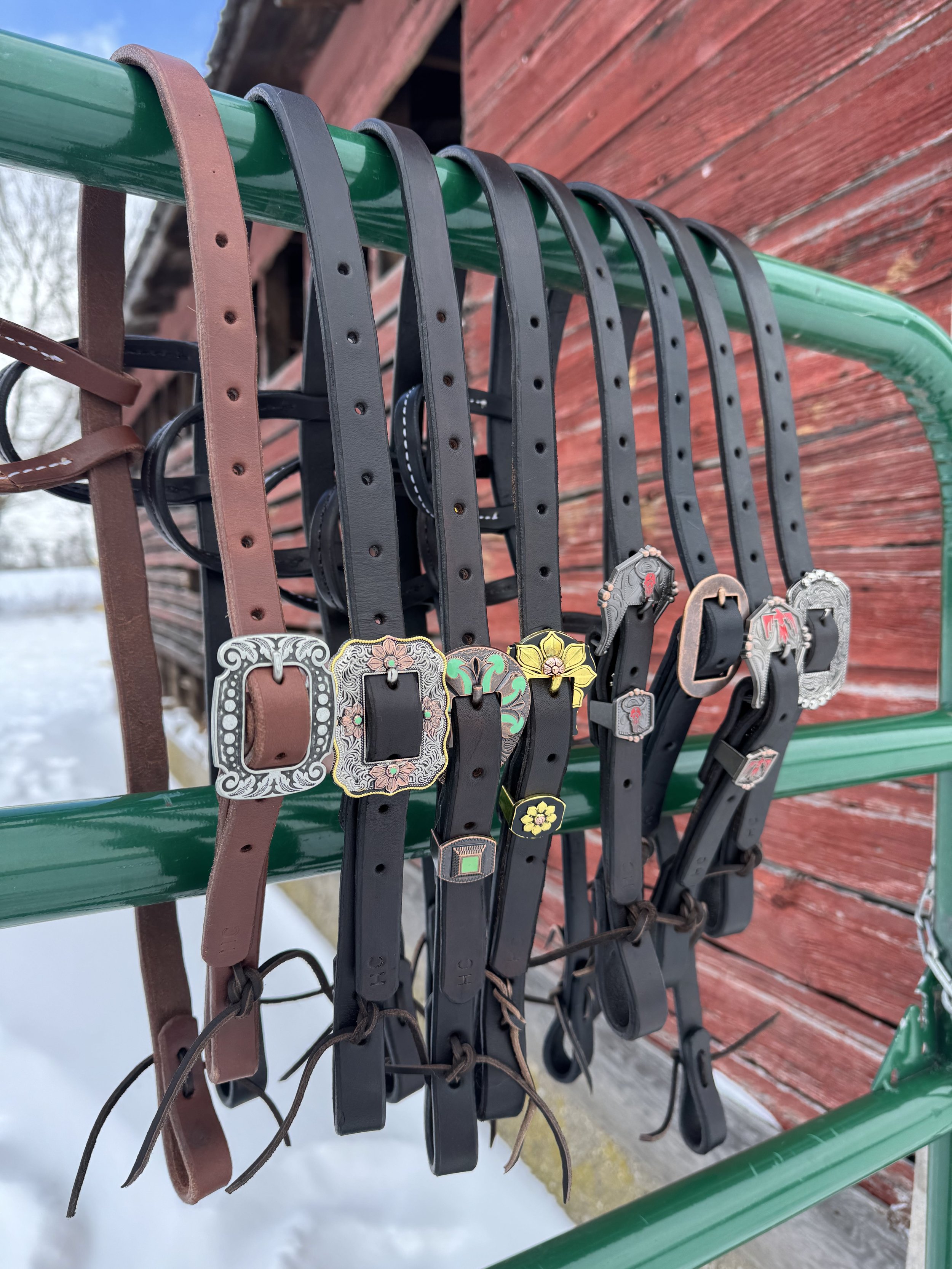 Six leather belts with metal buckles hanging on a green metal rack outdoors in front of a red wooden building, with snow on the ground.