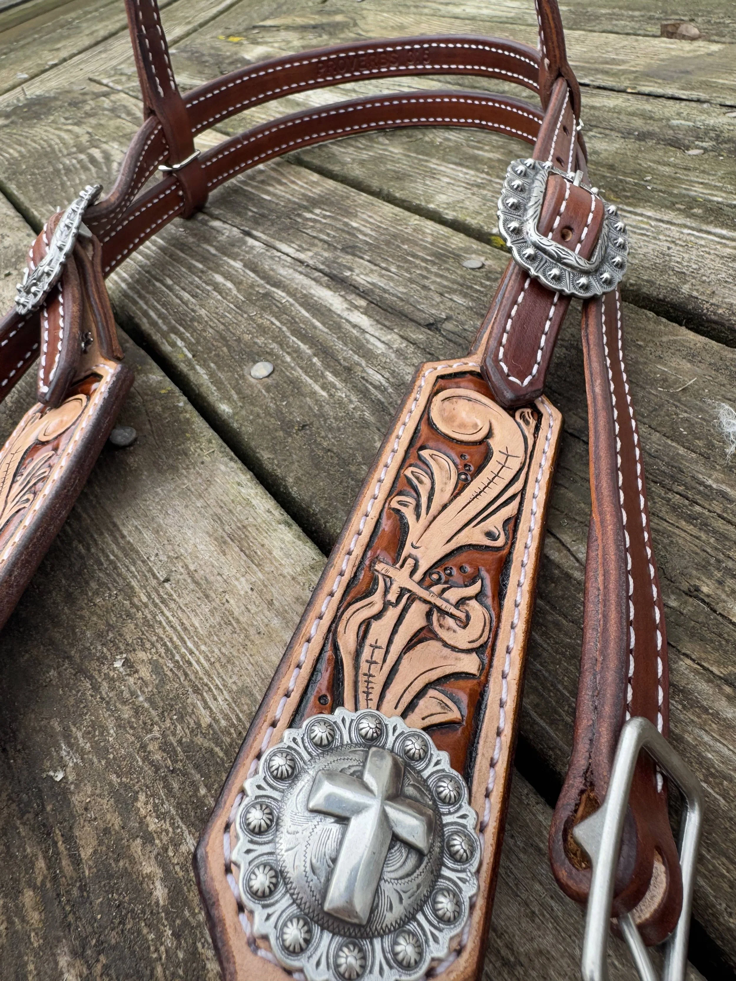 A close-up of a decorative western-style belt with a large silver buckle featuring an engraved skull and intricate floral patterns. The belt is made of leather with detailed tooling and white stitching, resting on a weathered wooden surface.