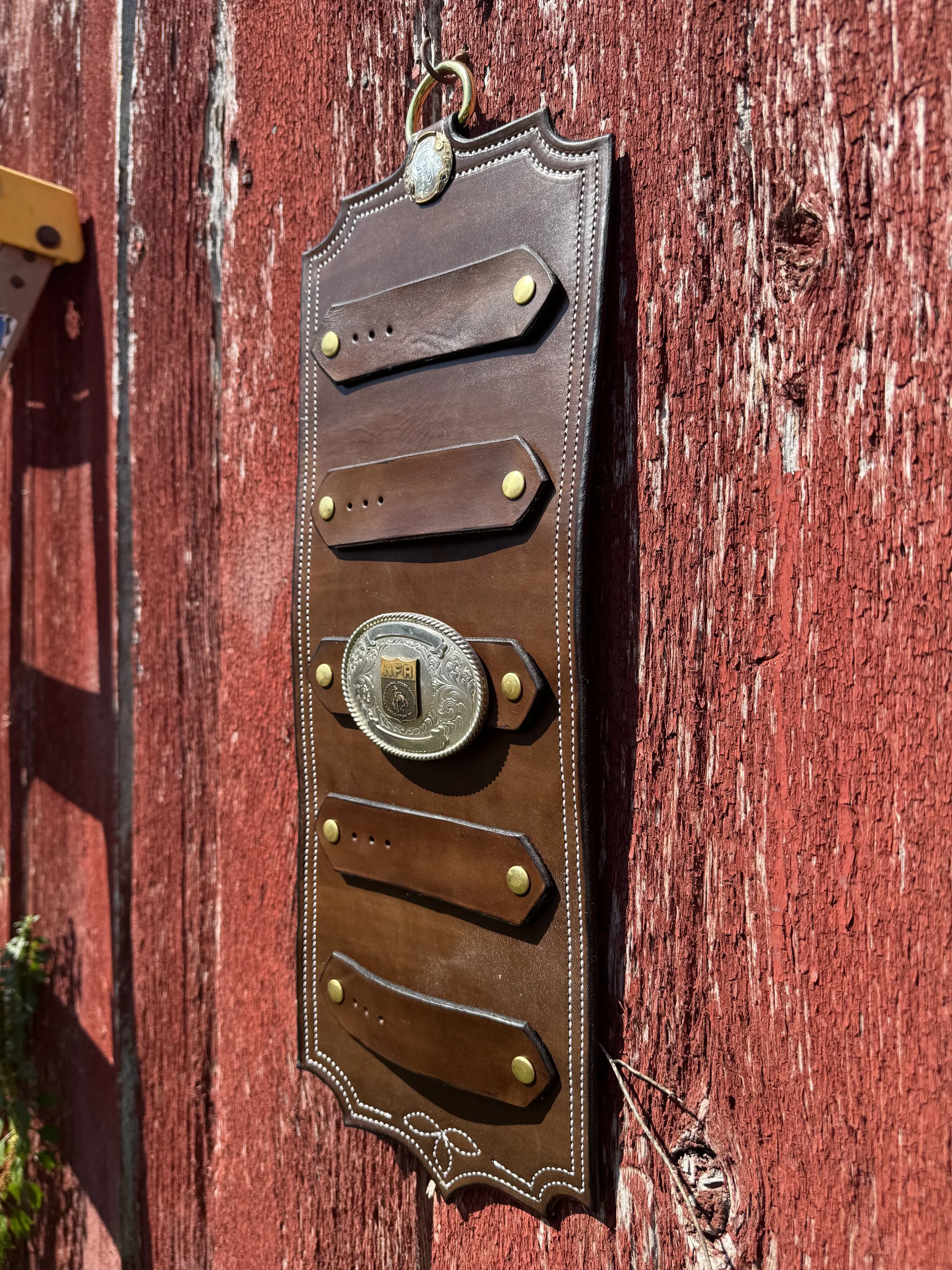 A decorative black and brown leather wall mount with brass accents and a silver embossed circle at the top, attached to a red, weathered wooden wall.