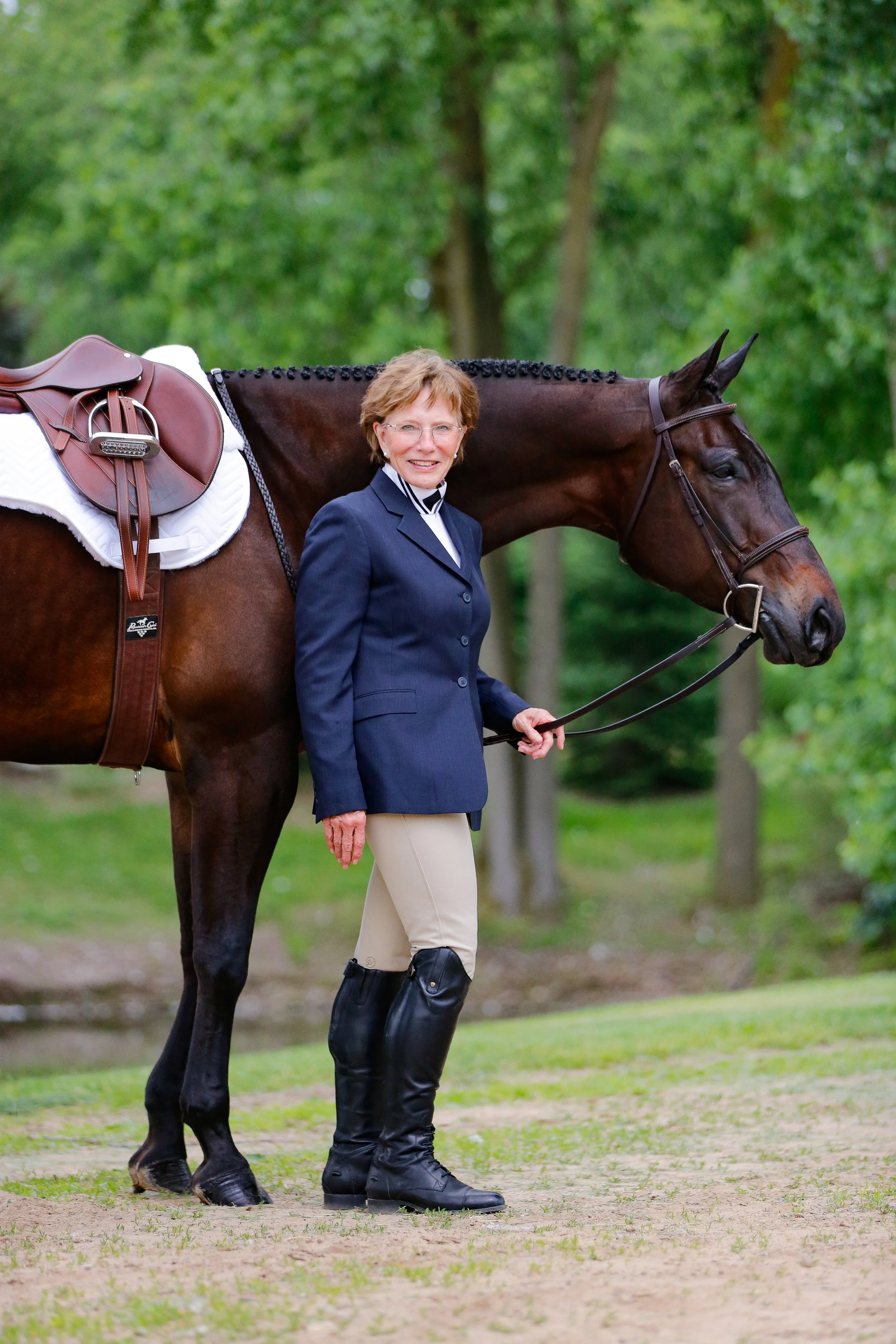 An older woman dressed in equestrian attire standing outdoors next to a brown horse carrying a saddle and bridle, with a background of green trees.