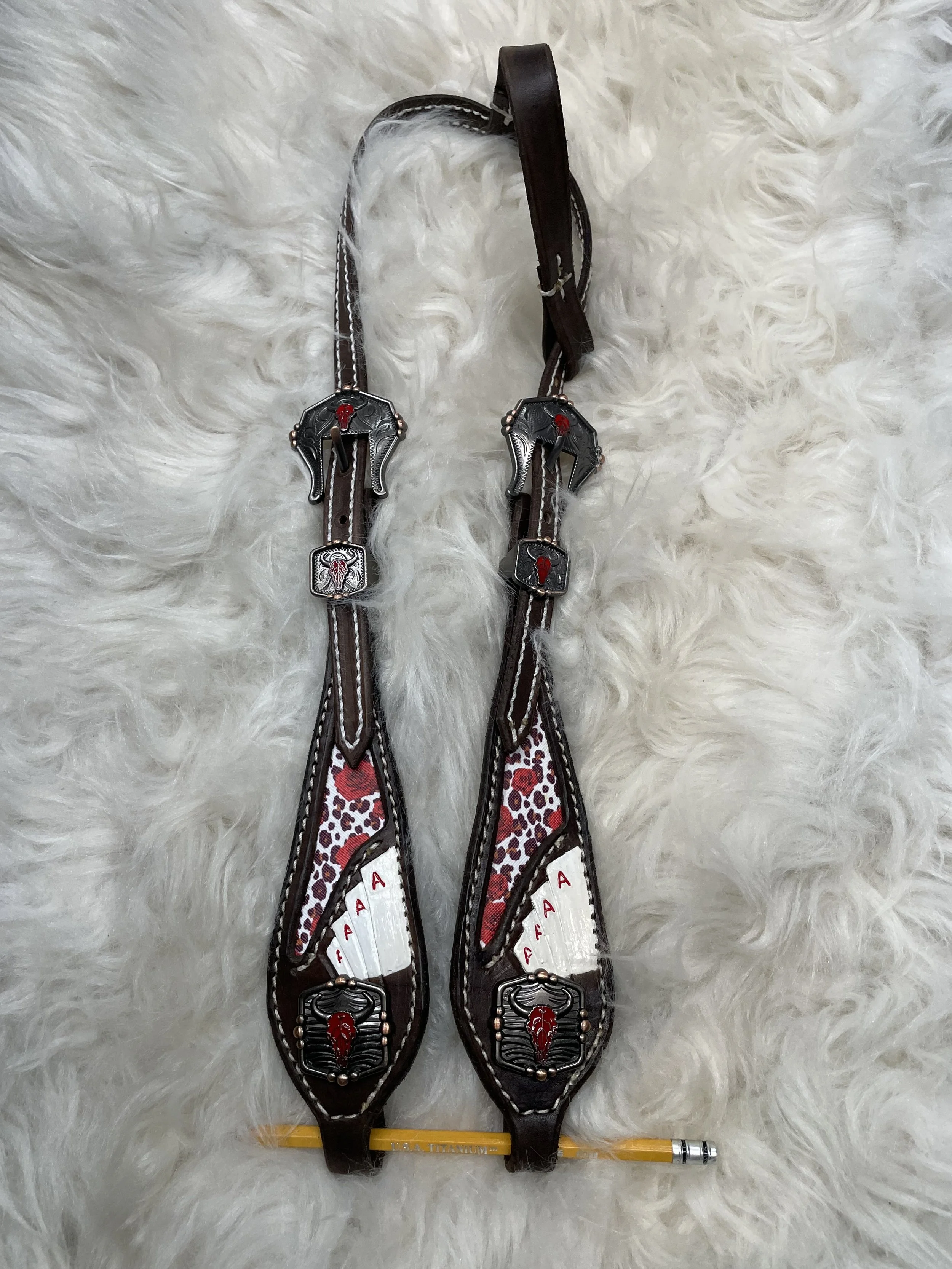 Pair of decorative boxing headgear with black straps, leopard print in red and white, silver embellishments, and an illustration of a head in a fierce pose on each piece, placed on a fluffy white surface along with a yellow pencil.