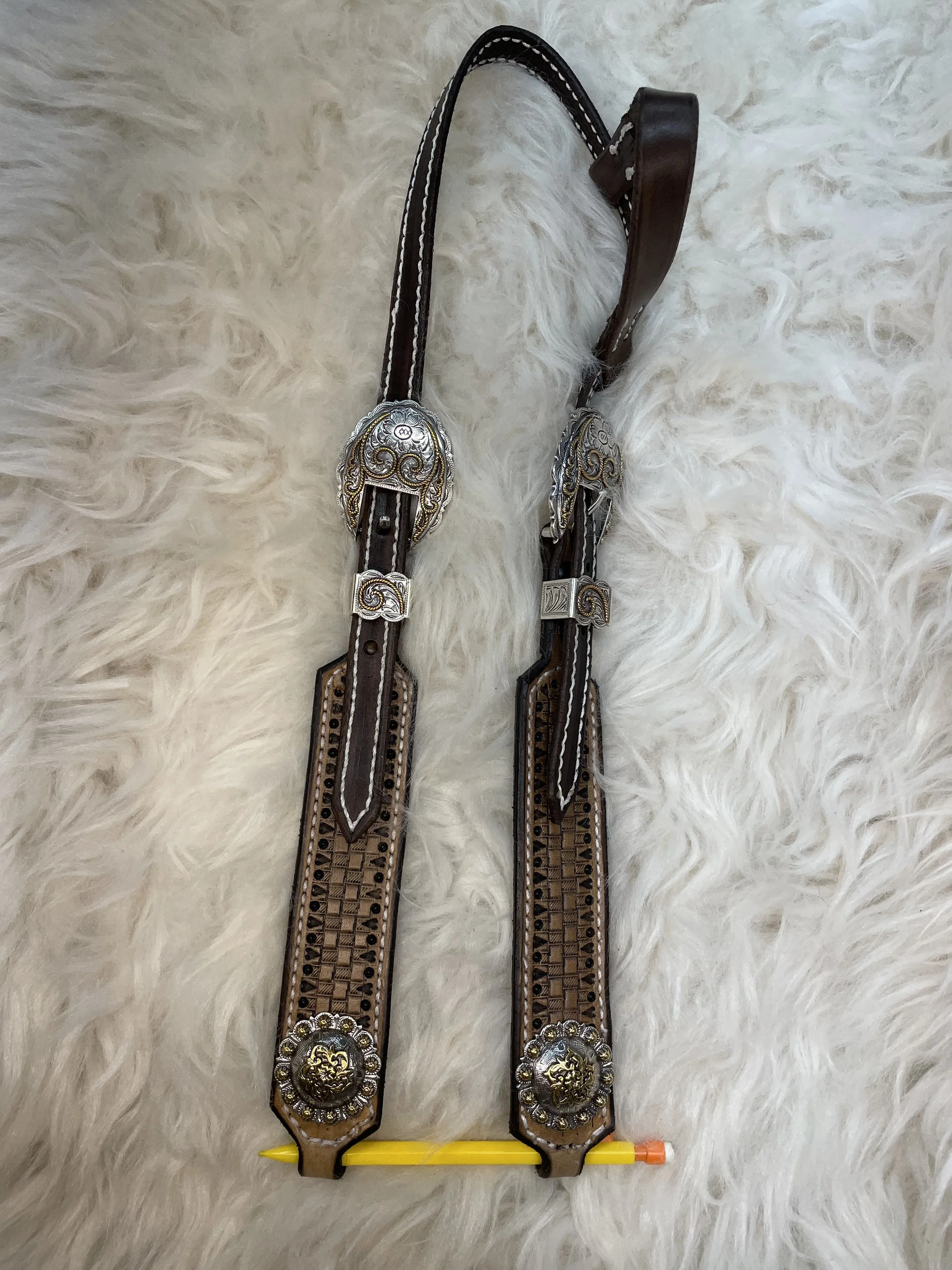 Decorative leather belt with ornate silver and gold embellishments lying on a white fluffy surface with a yellow and orange pen positioned underneath.