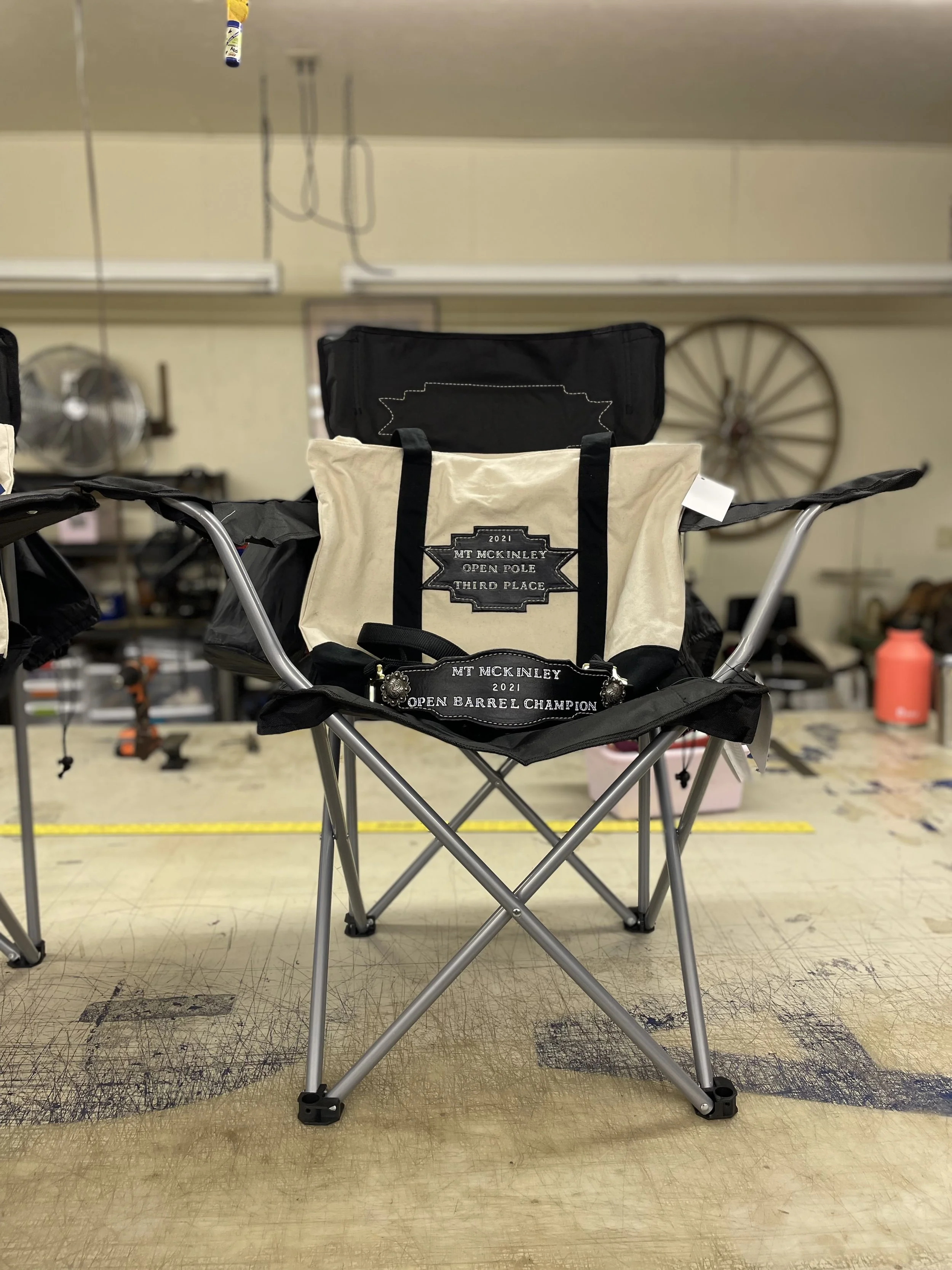 A desert camouflage folding chair in a workshop or storage room, with awards attached to the front, including a badge and fabric patch for 2021 Mount McKinley Open Barrel Champion and third place, respectively.