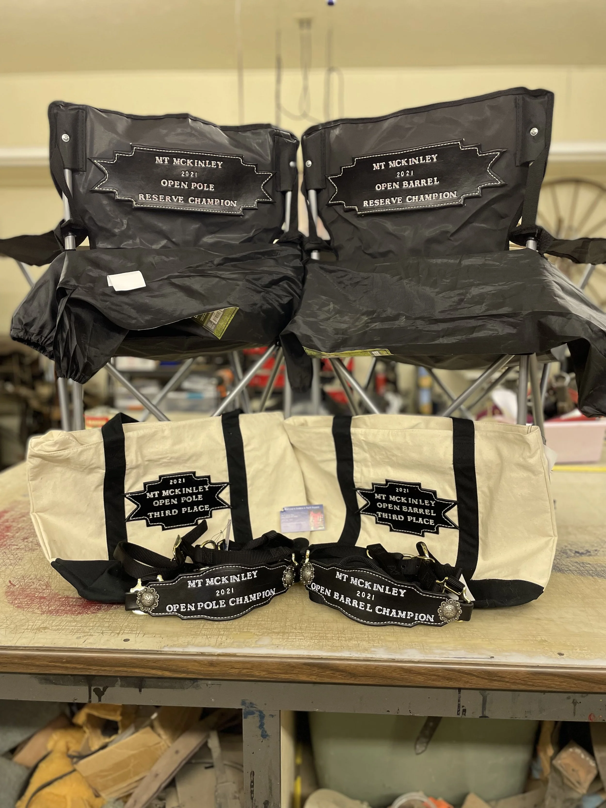 Display of awards and gear for a competitive event, including two large black bags with embroidered patches indicating '2021 Open Pole Reserve Champion' and '2021 Open Barrel Reserve Champion,' and smaller black wristbands with similar inscriptions a