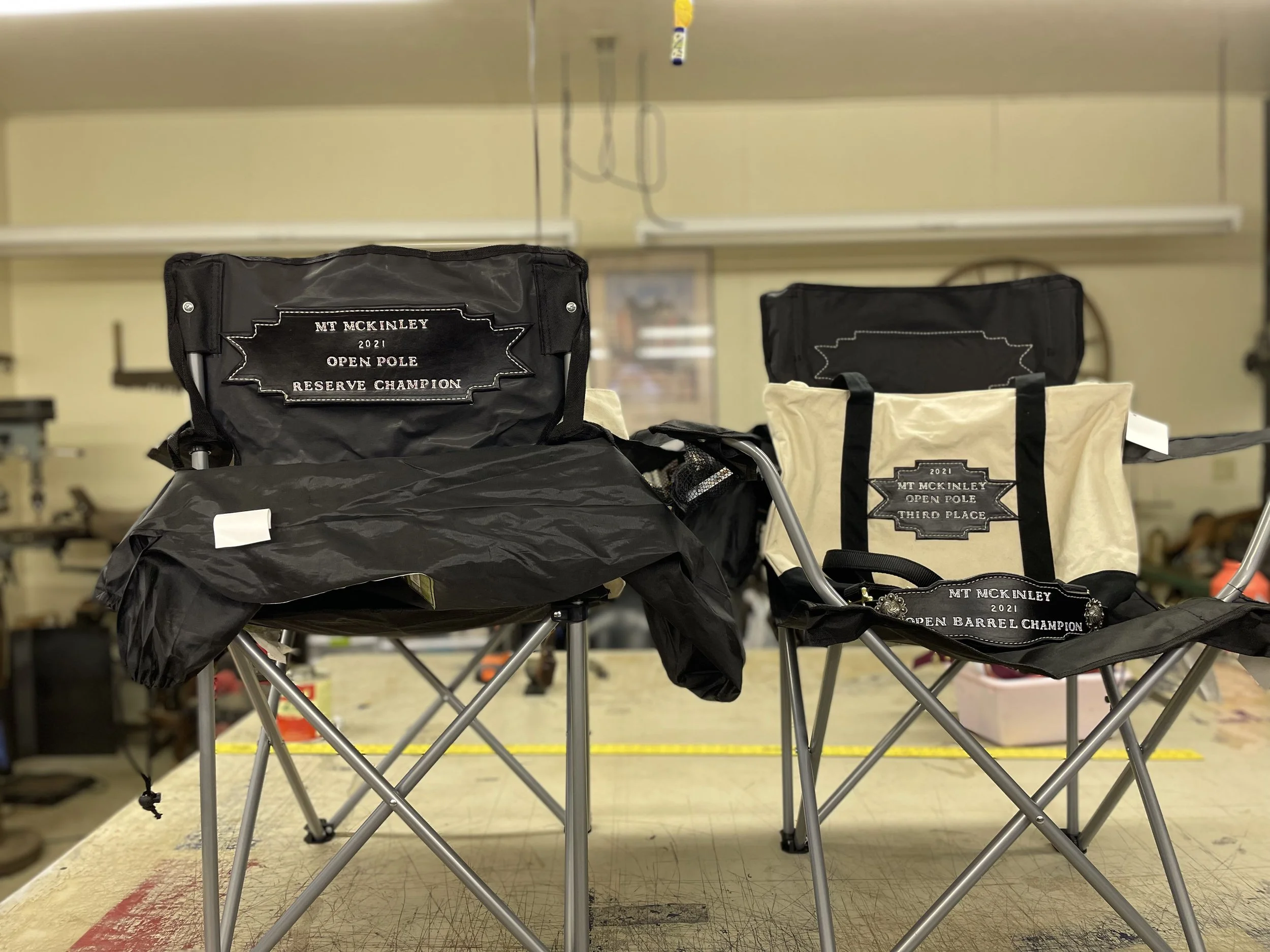 Black and beige tote bags with embroidered patches reading '2021 Open Pole Reserve Champion' and '2021 Open Barrel Champion' are placed on a table in a room with beige walls and equipment.