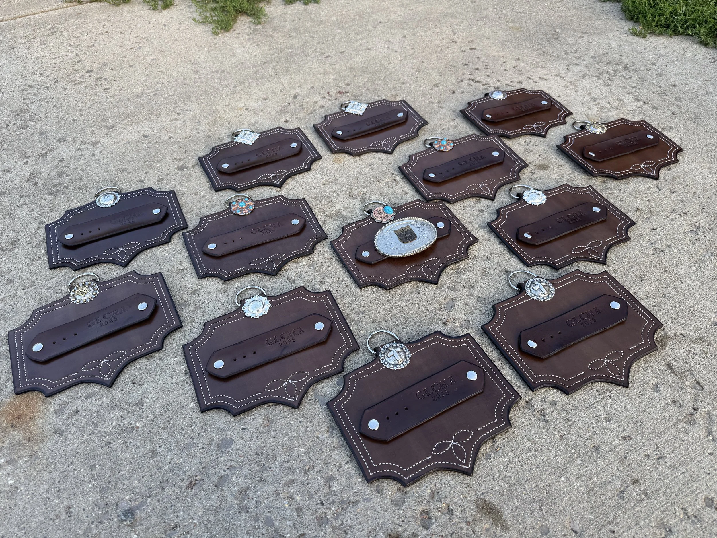Multiple decorative leather tag plaques with silver accents and jewelry, laid out on a concrete surface with some green shrubbery at the top.