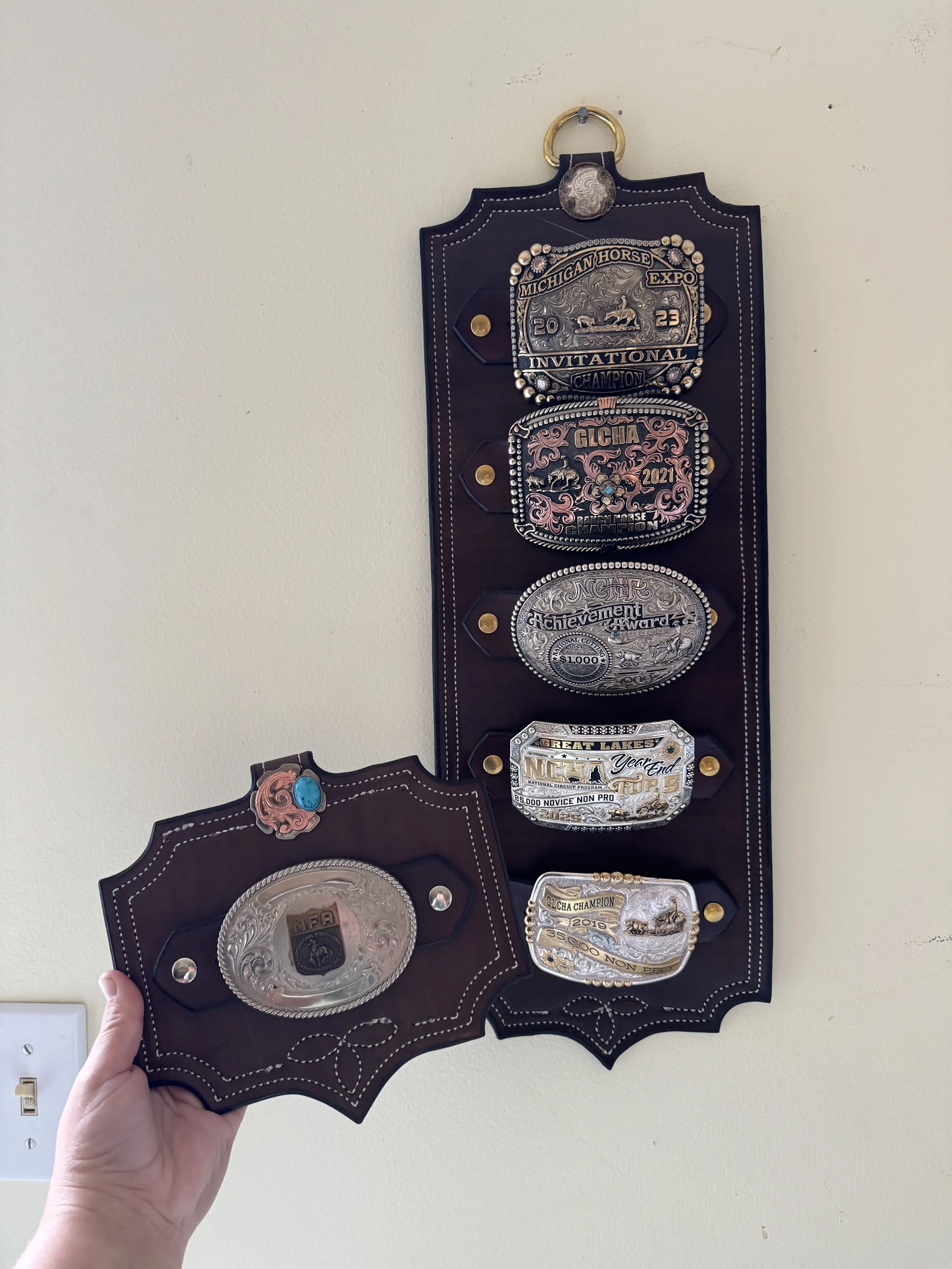 A person holding a wooden plaque with multiple large silver and gold belt buckles and awards attached to it, displayed on a wall.