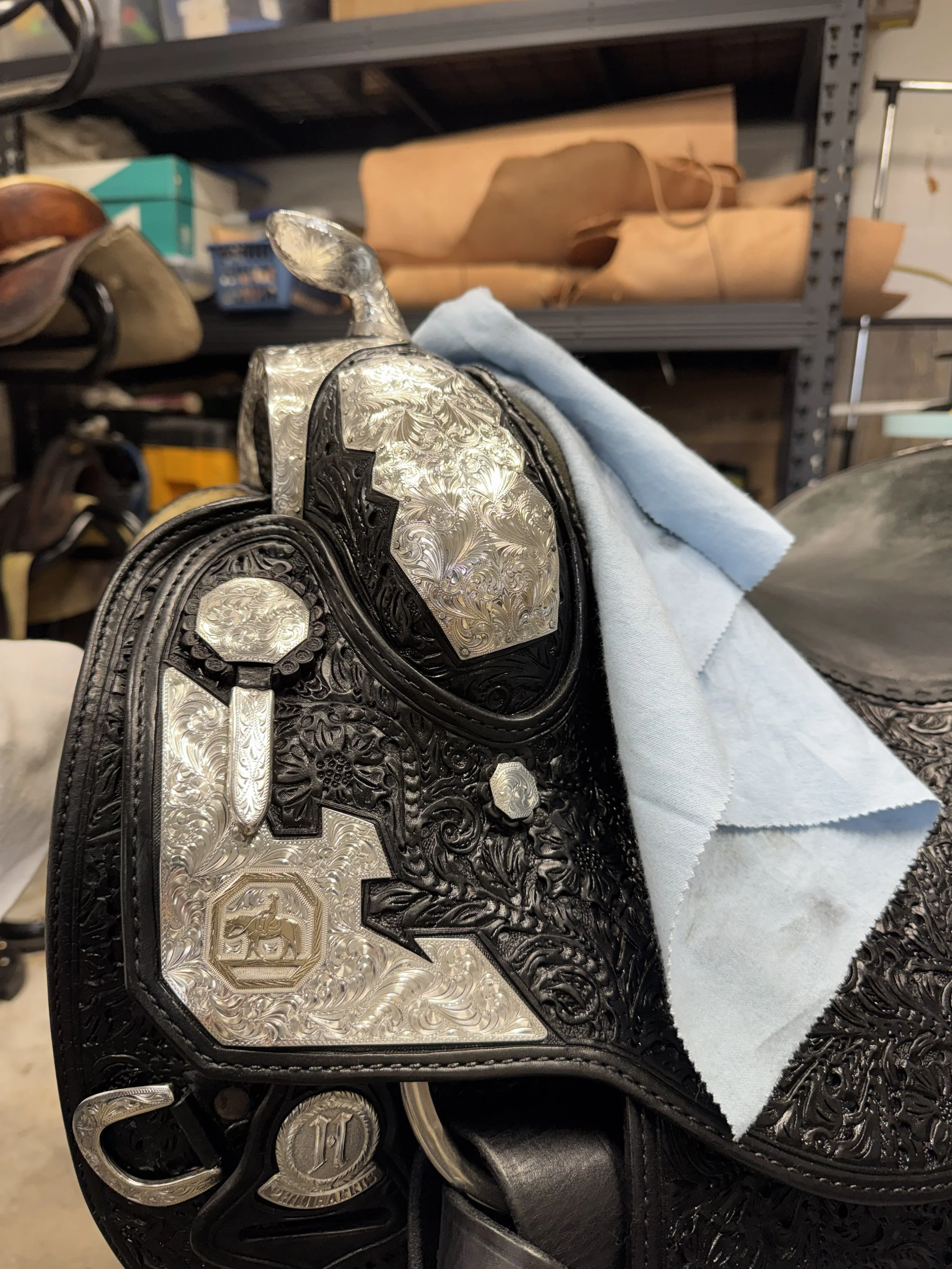 Close-up of a shiny, engraved silver belt buckle featuring a 10-cent coin, with a white towel draped over a black leather item, set against a background of shelves with various leather goods and materials.