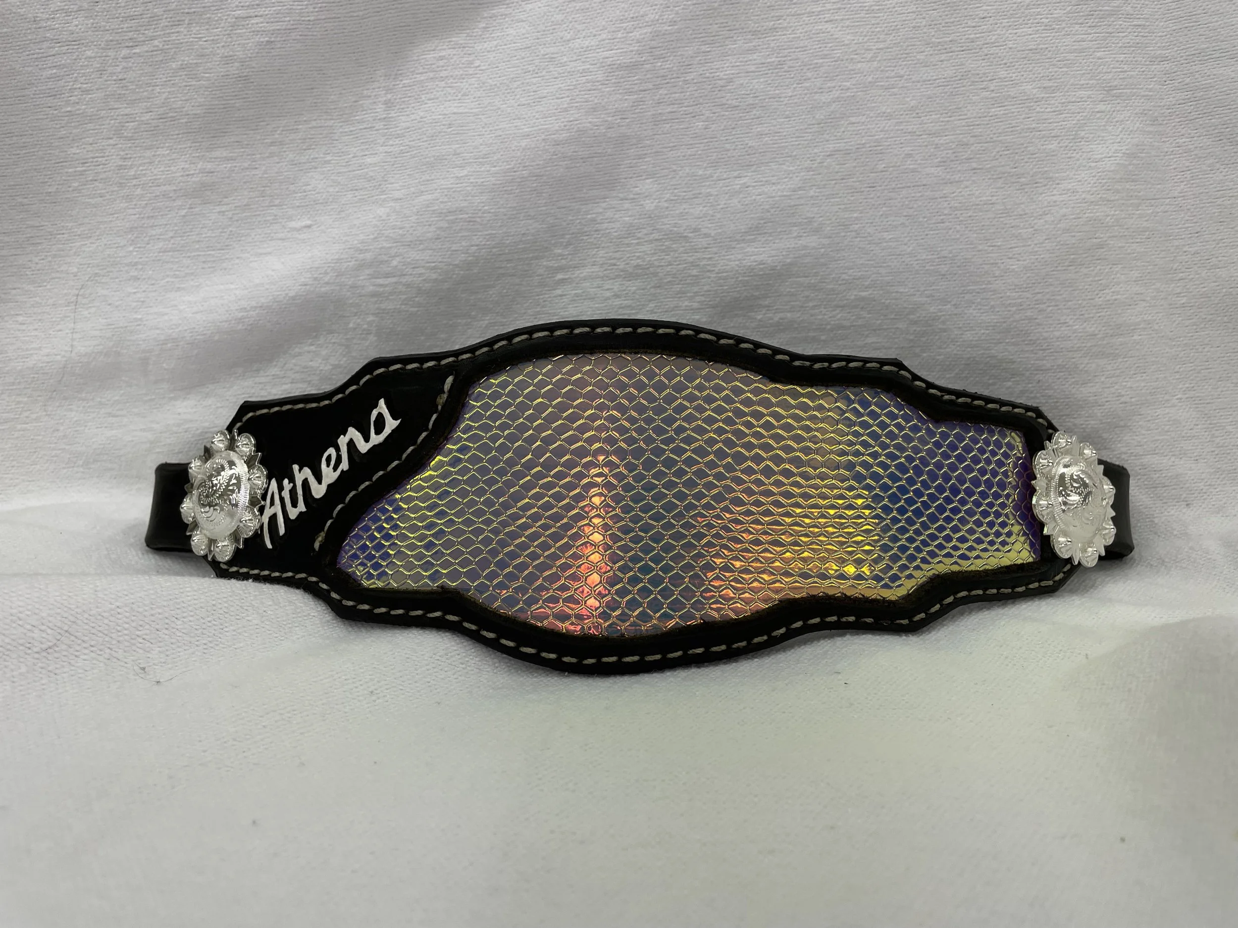 A decorative accessory or badge with holographic, reflective surface and ornate silver embellishments on each side, placed on a light gray fabric background.