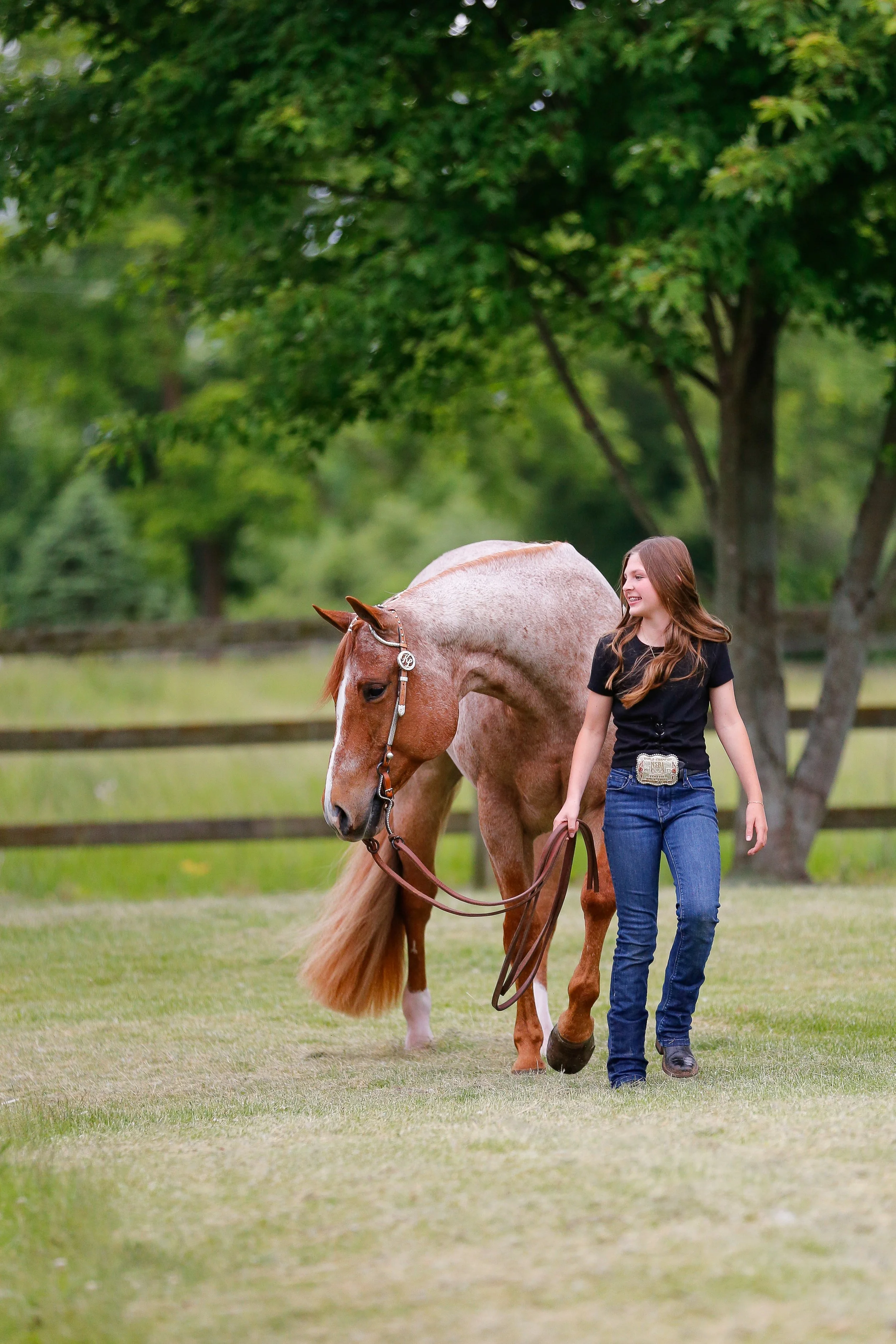 A young woman with long brown hair walking a light brown palomino horse on a grassy field with trees in the background. The woman is smiling and holding the horse's reins.