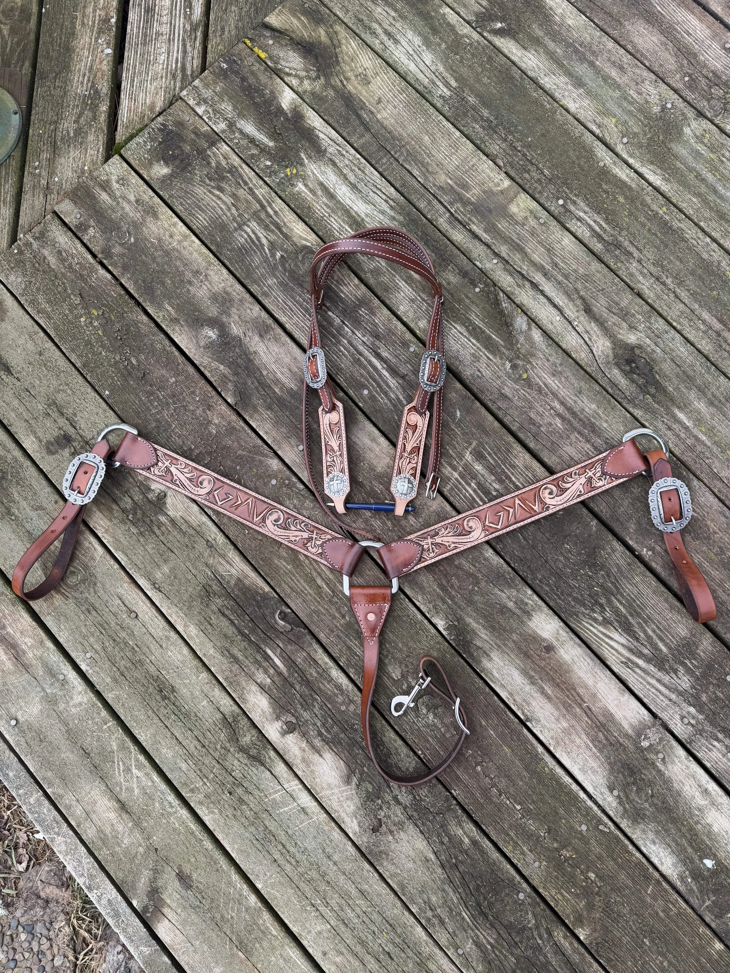 A leather western-style horse bridle with decorative silver accents, laid out on a weathered wooden deck with a diagonal pattern.