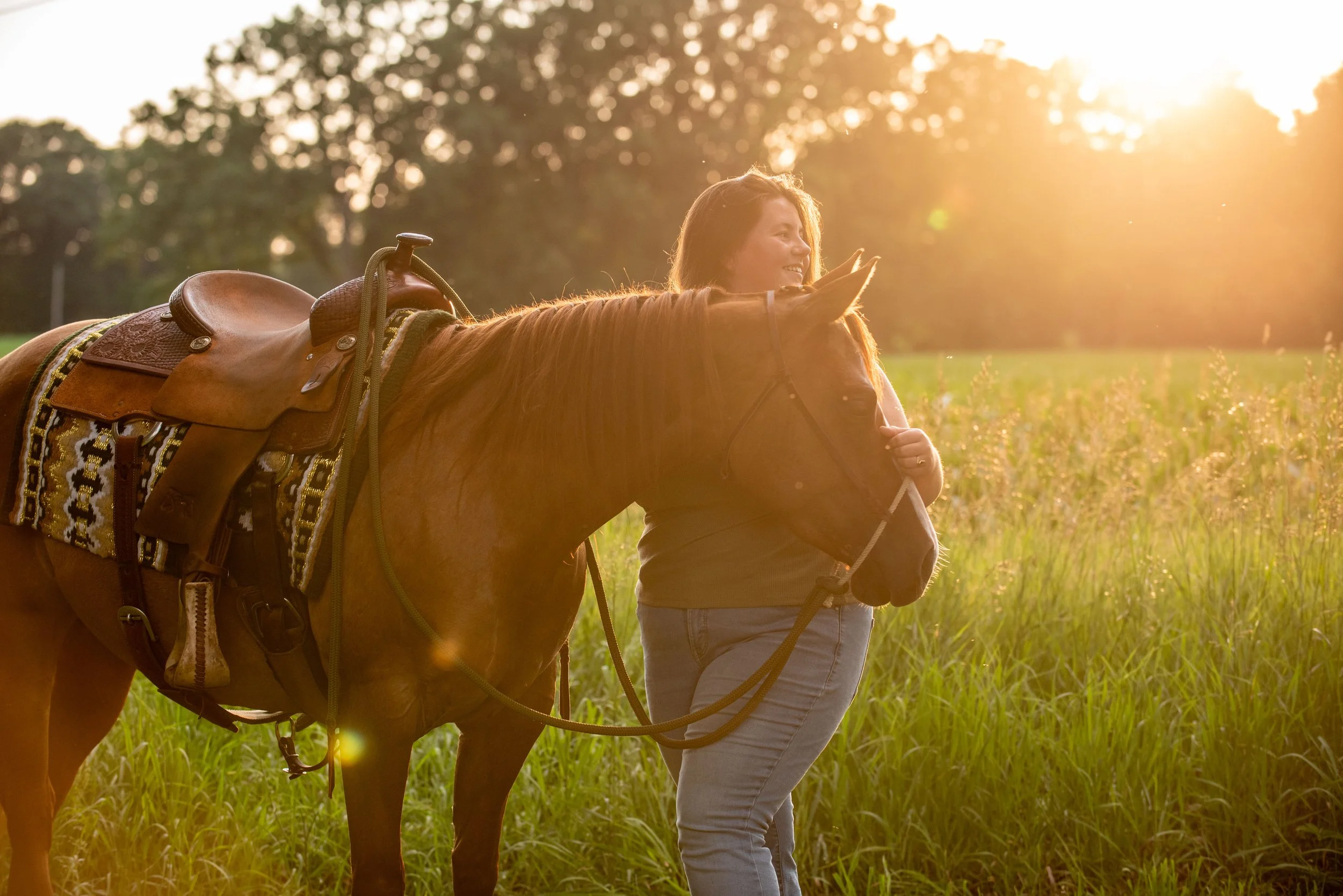 A woman smiling and holding a young horse's reins in a sunlit field at sunset.
