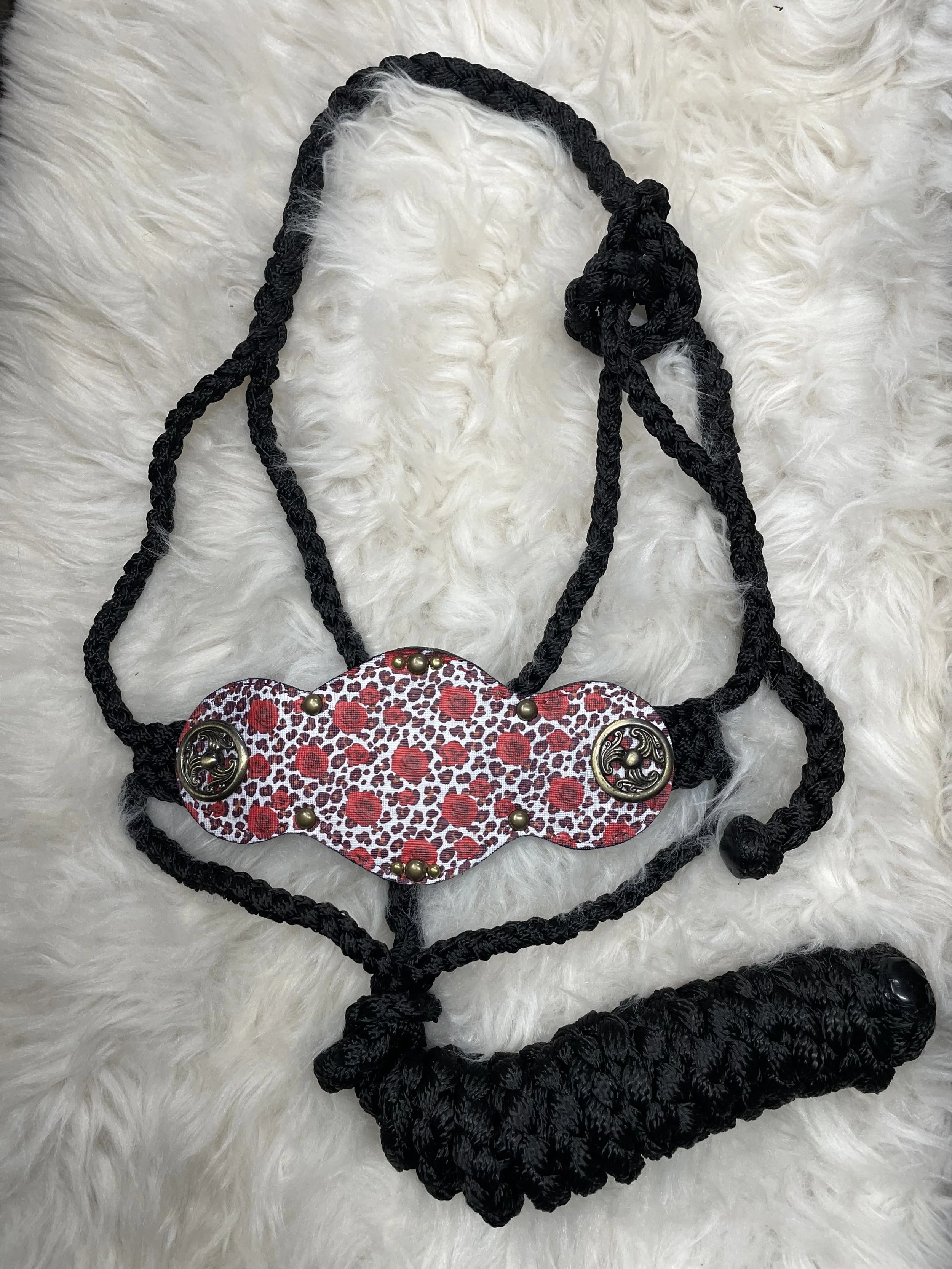 A black braided rope necklace with a decorative fabric piece featuring a red and white animal print pattern and metallic accents, laid on a white fluffy surface.