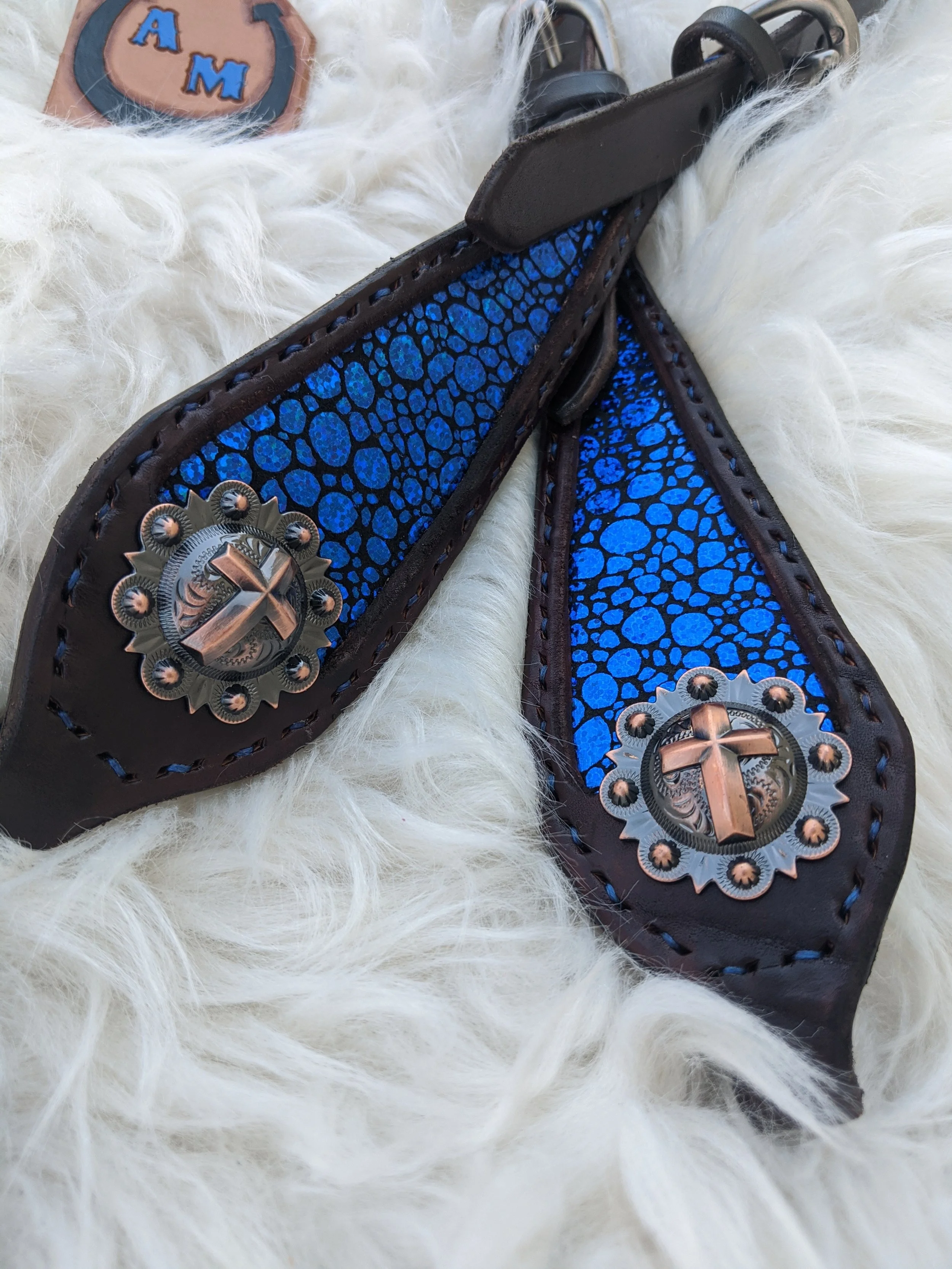 Close-up of a pair of black cowboy boots with blue and black textured vamp, silver and copper decorative conchos, and white furry background. Part of a patch with the letters 'A' and 'M' is visible.