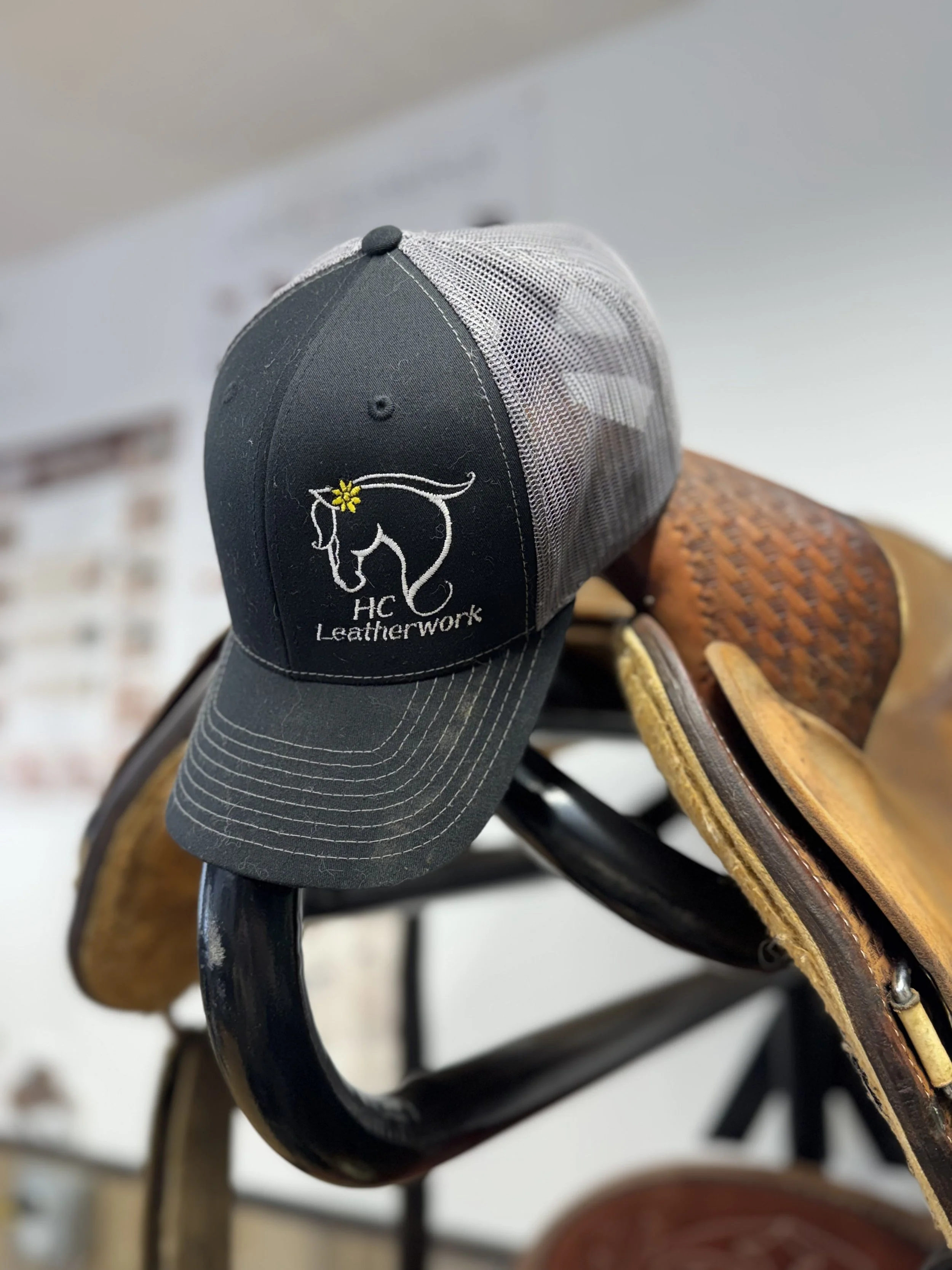 Close-up image of a black baseball cap with a horse logo and the text 'HC Leatherwork' embroidered on the front, resting on a wooden saddle on a cart or wagon.