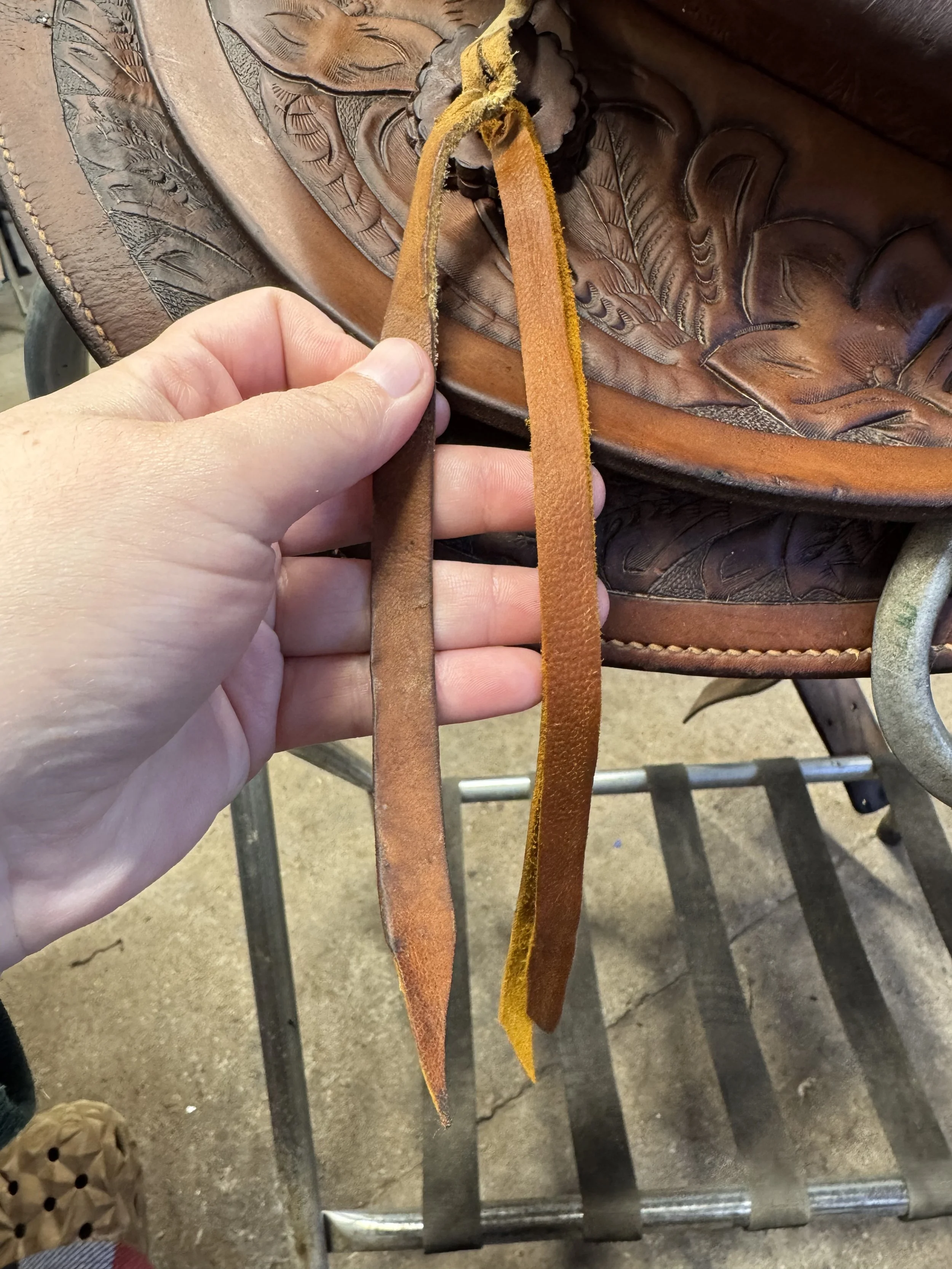 A person holding thin leather strips in front of a carved leather saddle with detailed floral and leaf patterns.