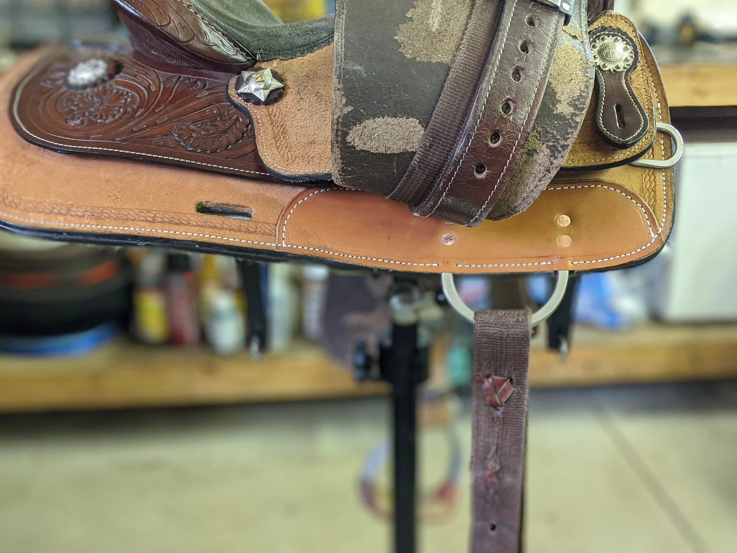 Close-up of worn leather belts, including a cowboy belt with decorative stitching and a metal buckle, draped over a saddle or similar equipment with a background of a workshop or store.