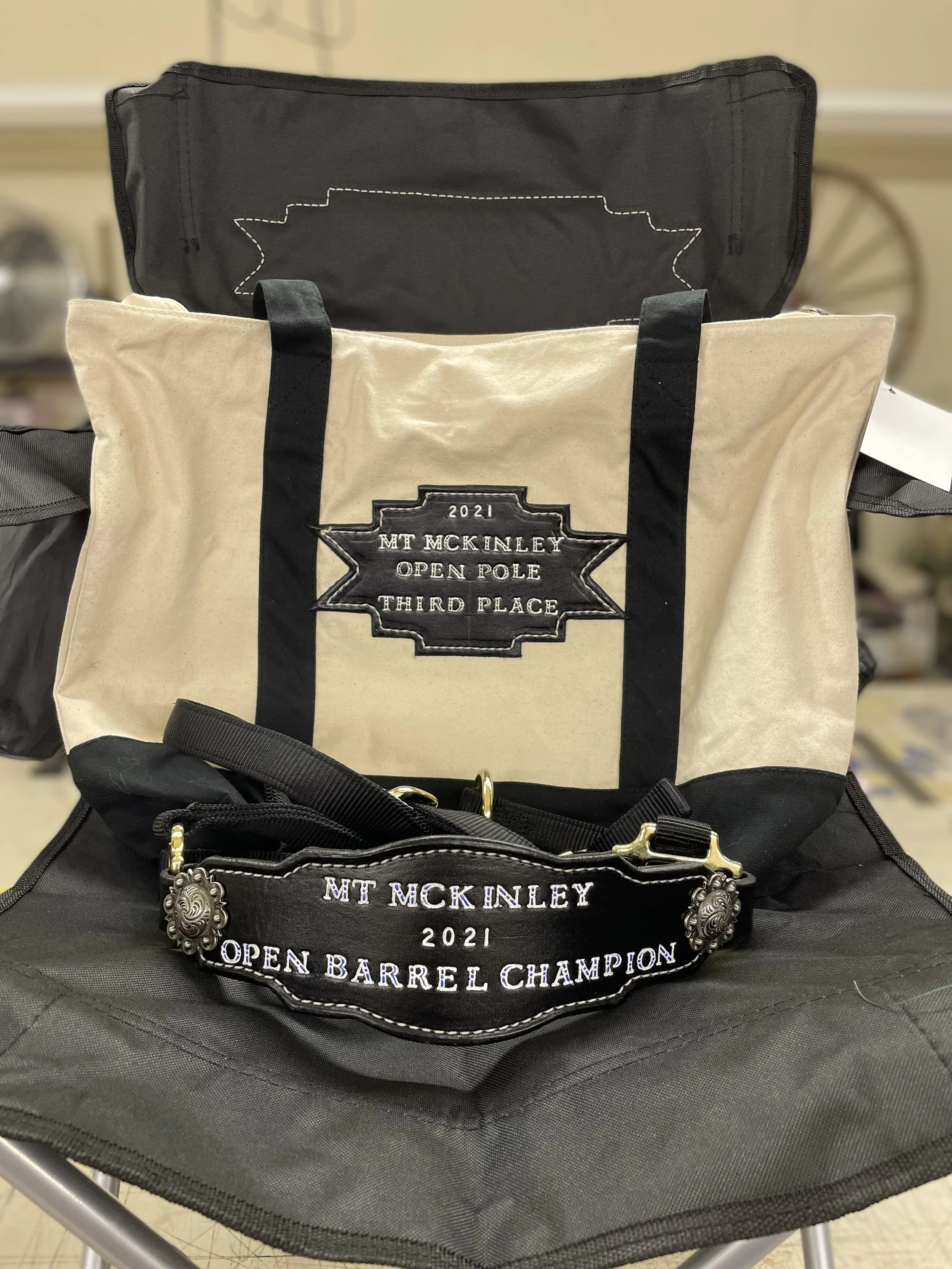 A beige tote bag with black handles and a patch that reads '2021 MT MCKINLEY OPEN BARREL CHAMPION THIRD PLACE' is placed inside a larger black bag with a metal clip attached to the front.