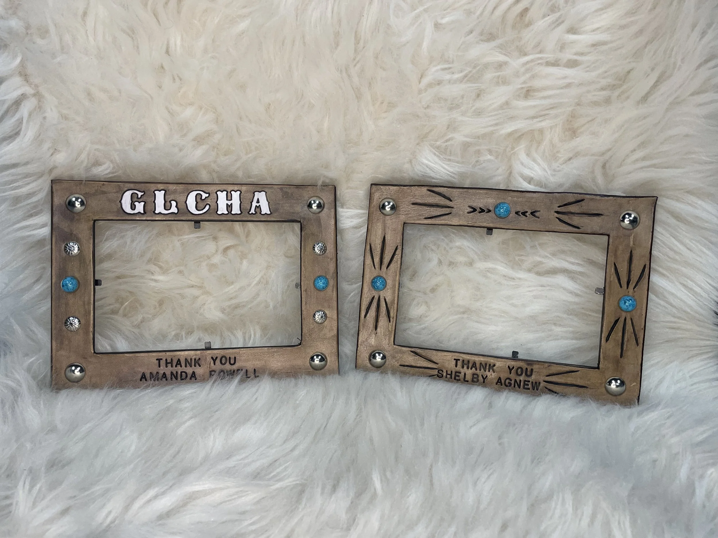 Two decorative wooden frames with blue and silver embellishments, placed on a fluffy white surface. The left frame says "G.L.C.H.A" and has a note underneath that reads "Thank you Amanda Powell." The right frame says "Thank you Shelby Agnew." Both fr