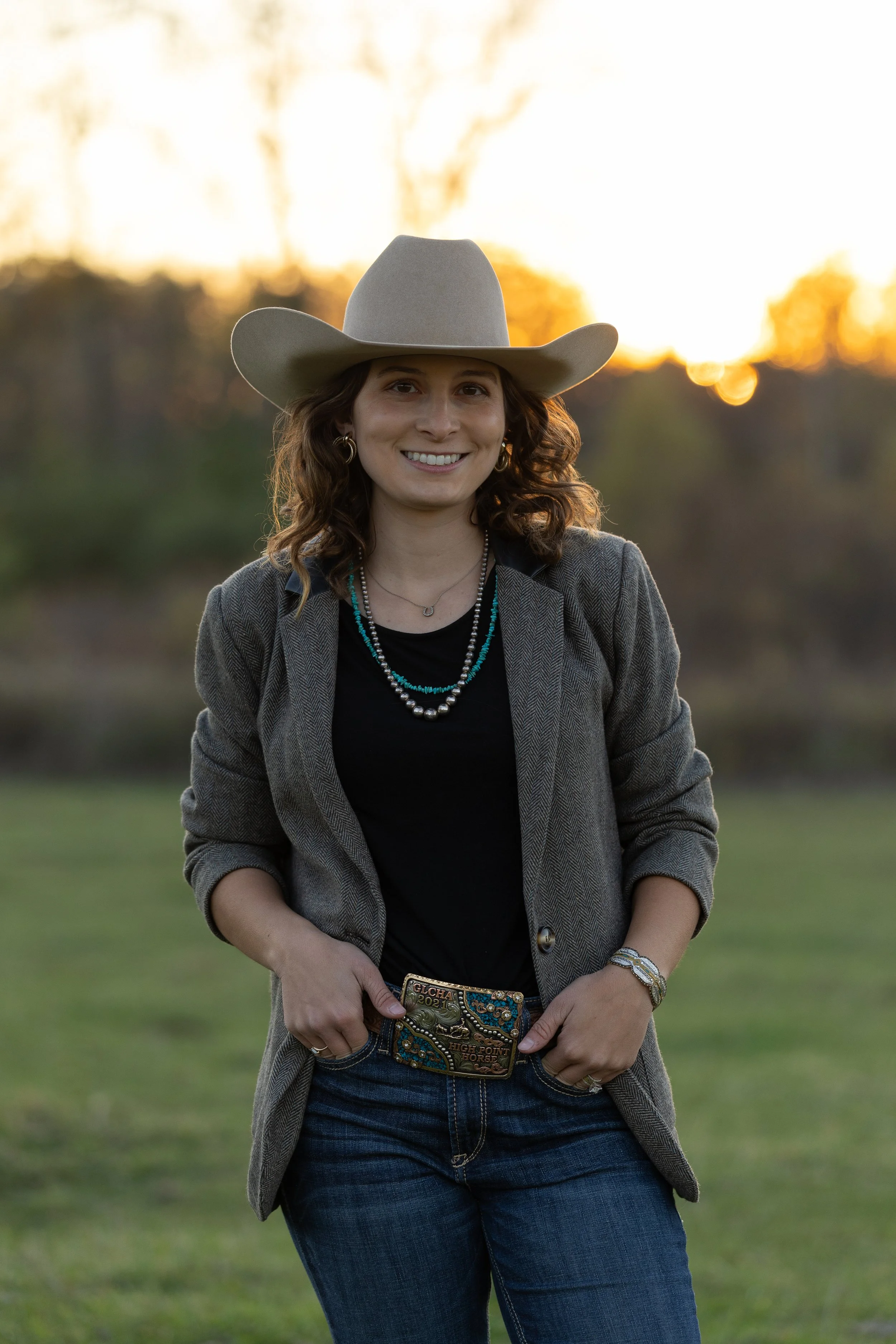 Smiling woman wearing a cowboy hat, jewelry, blazer, and jeans standing outdoors at sunset.